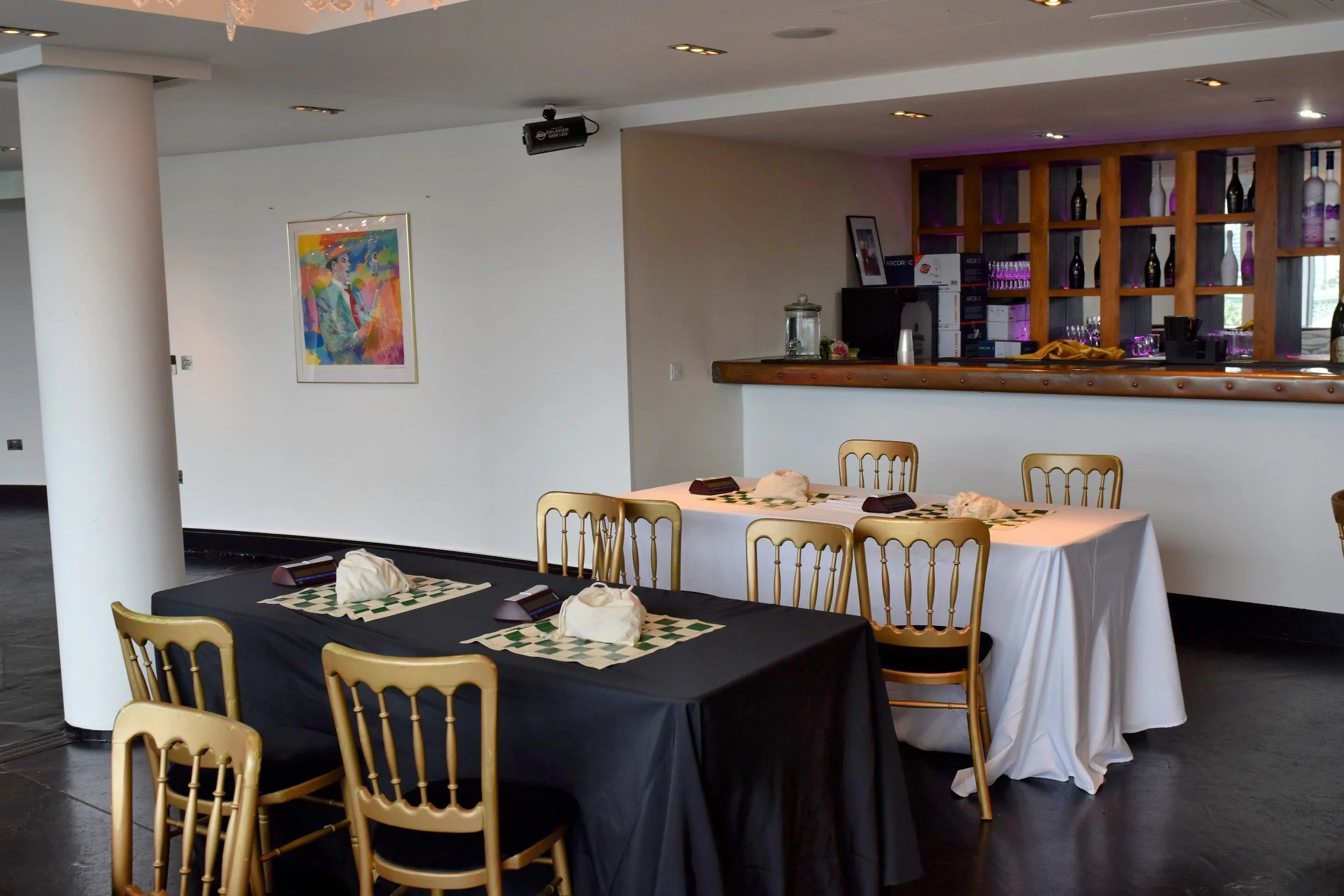 Interior of a restaurant or event space with tables set for dining, including napkins, plates, and glasses, with a colorful framed painting on the white wall and a bar area with shelves holding bottles and glasses.