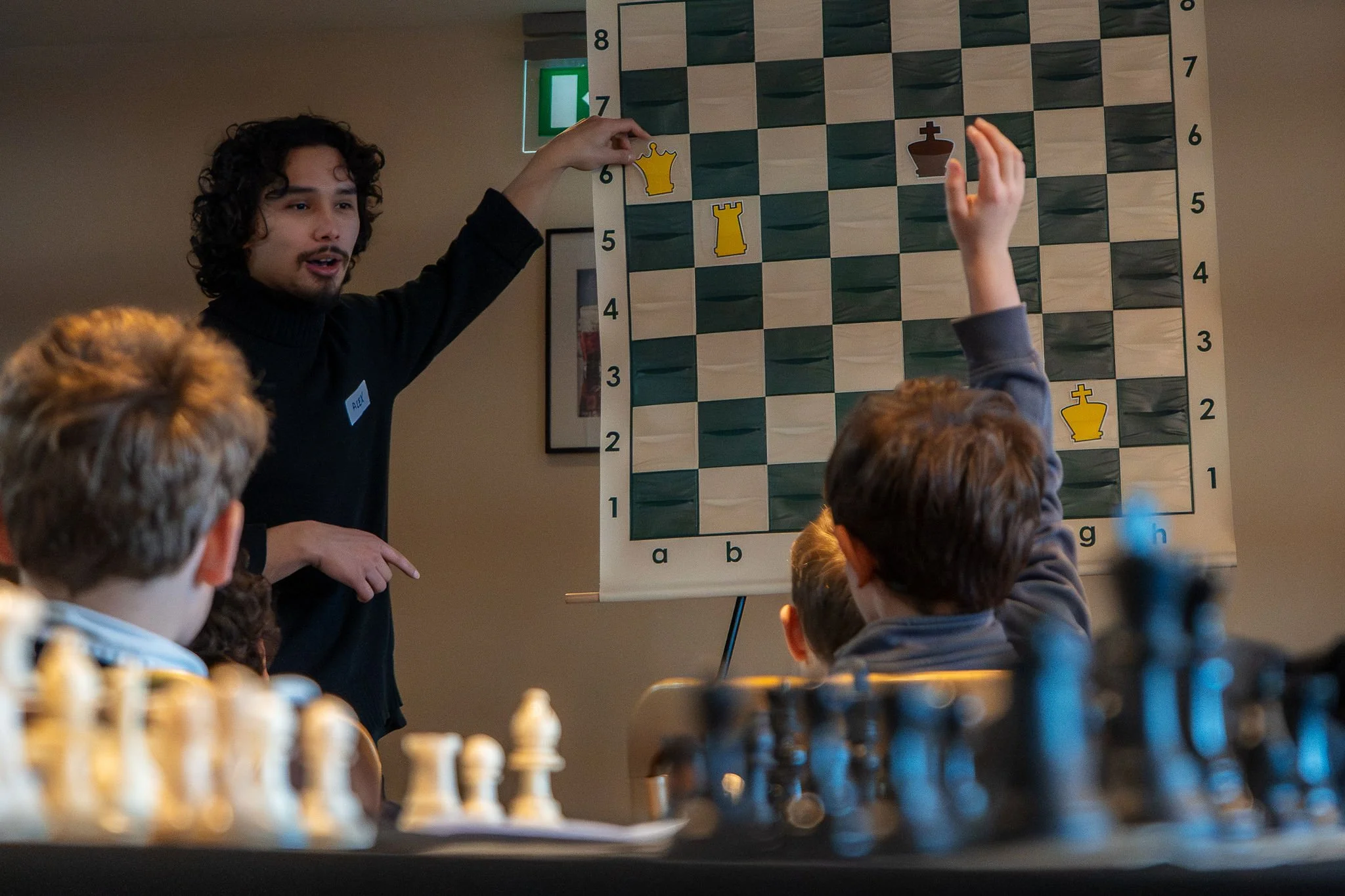 A man with curly hair and a beard giving a chess lesson to children. He is standing next to a large chessboard on a wall, with children seated at a table in the foreground, some raising their hands, inside a room with beige walls.