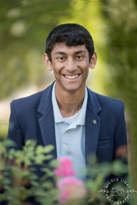 Teen boy smiling outdoors in a park, wearing a blue blazer and a white shirt, with blurred green trees in the background.