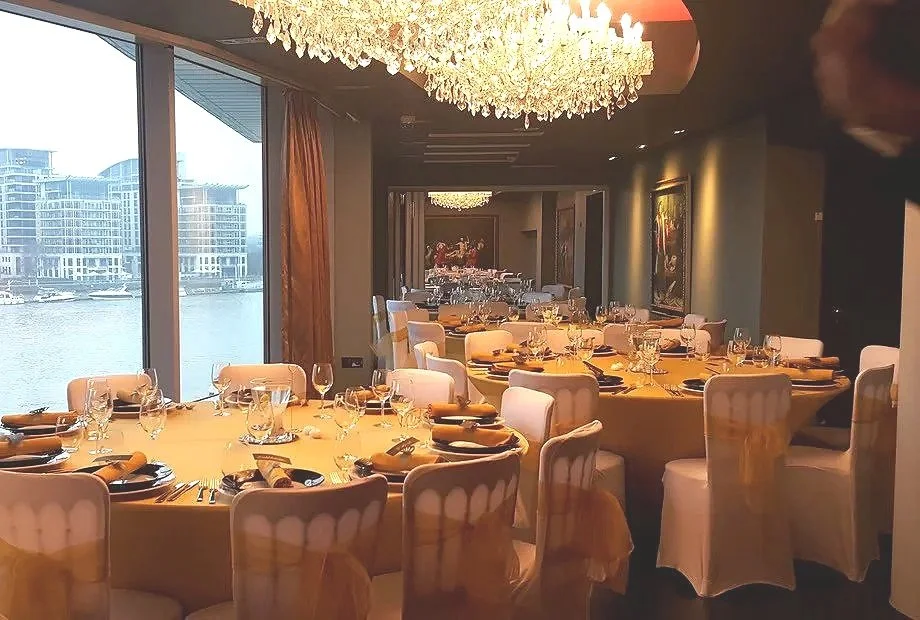 Elegant dining room set for a formal event with a chandelier, round tables covered with yellow tablecloths, and chairs with white covers. Large windows show a waterfront cityscape in the background.