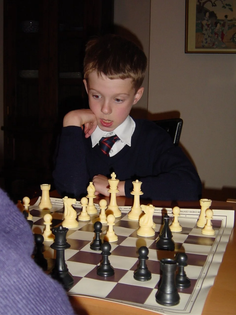 A young boy with brown hair and wearing a school uniform, including a white shirt, dark sweater, and a plaid tie, is playing chess. He is sitting at a table with a chessboard in front of him, focusing on the game.