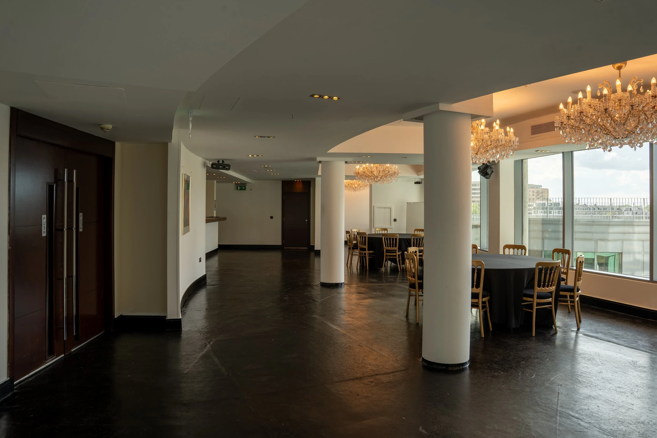 A spacious banquet hall with large windows, chandeliers, round tables with black tablecloths, and wooden chairs, featuring white columns and a view of city buildings outside.