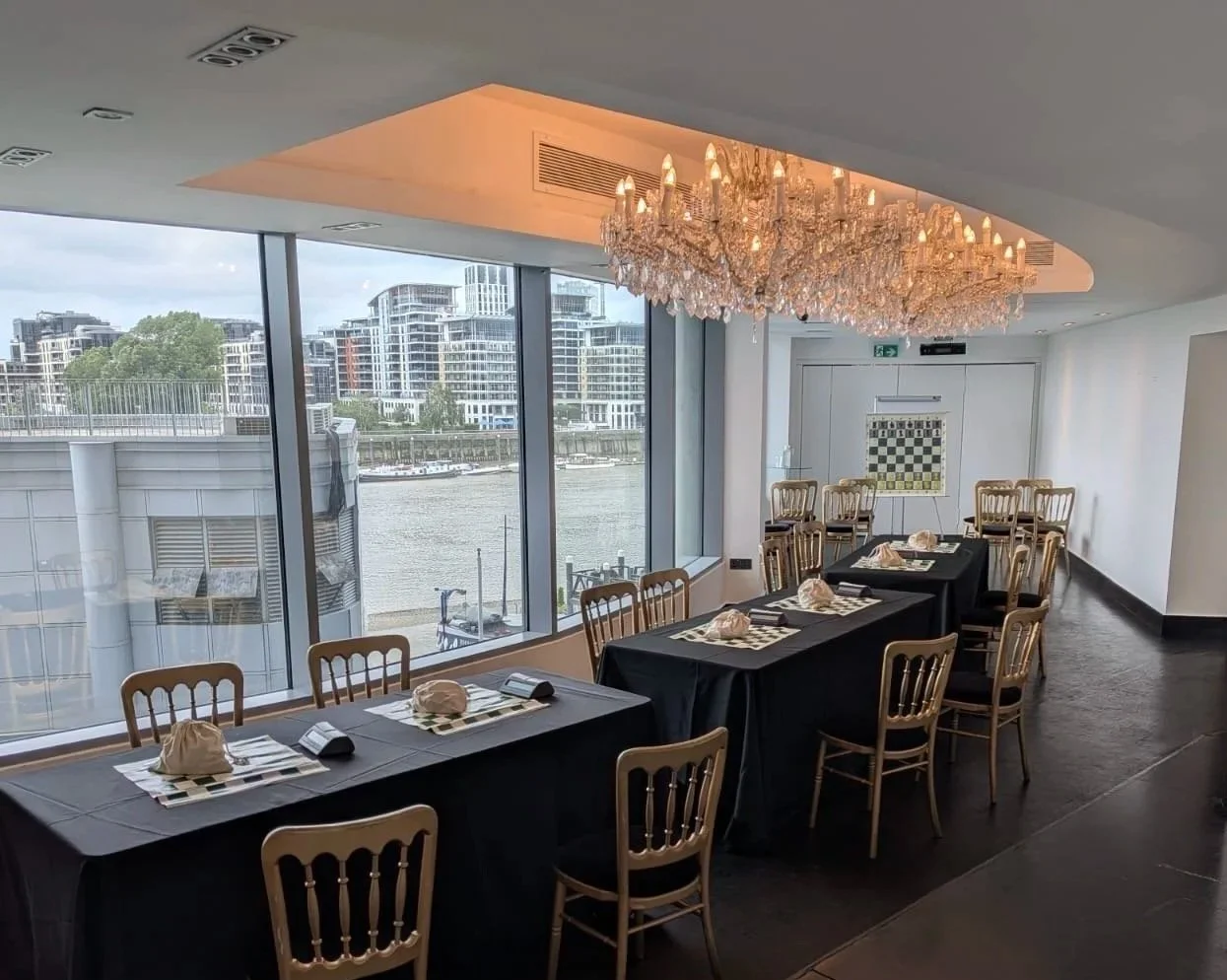 Elegant dining room with black tablecloth-covered tables, set with napkins and checkered placemats, gold chairs, large chandelier, and large windows overlooking a river and modern buildings.