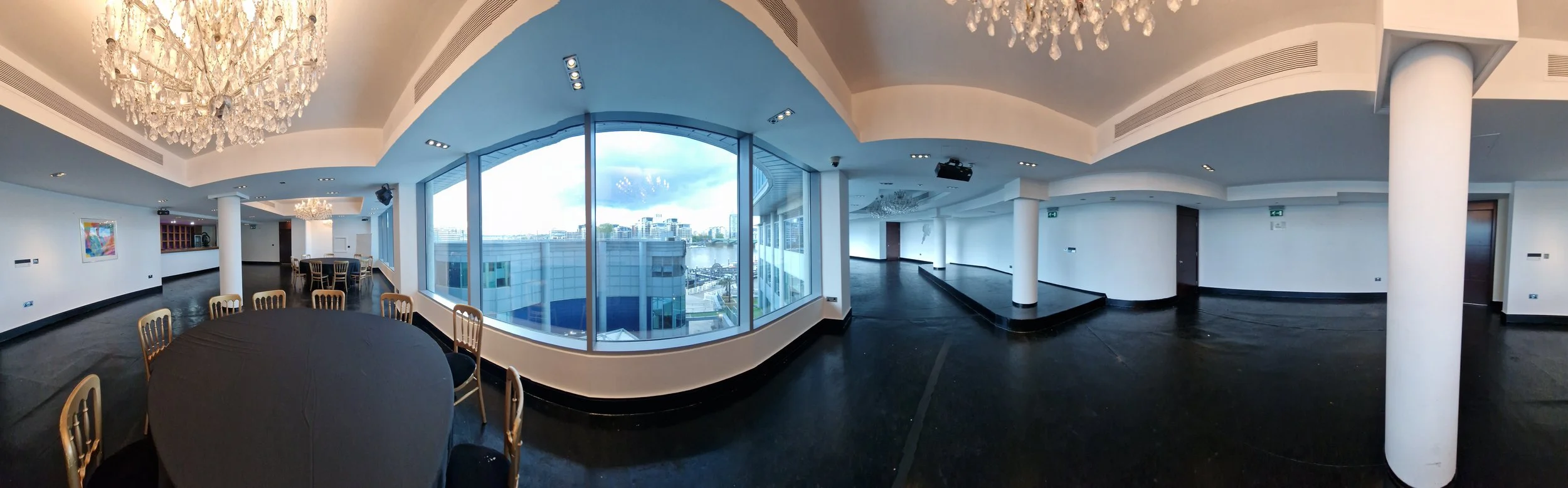 Spacious, modern event room with large windows, a chandelier, round table with chairs, artwork on the wall, and city view outside.