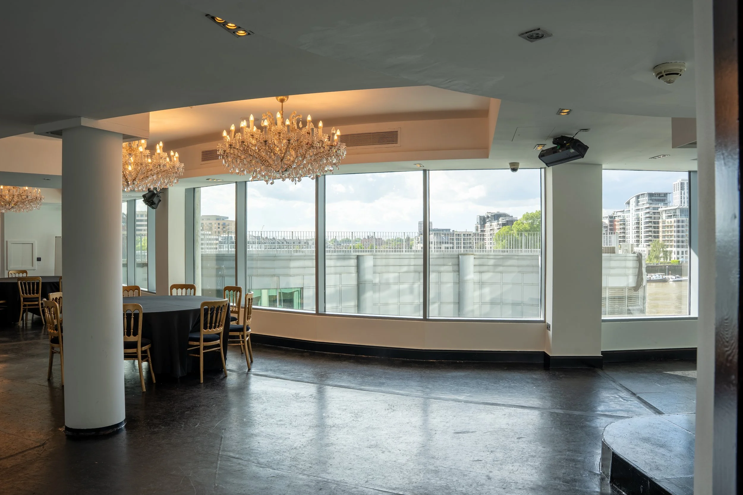 Interior of a modern event space or restaurant with large windows showing city buildings outside, decorated with crystal chandeliers and black-draped tables surrounded by wooden chairs.