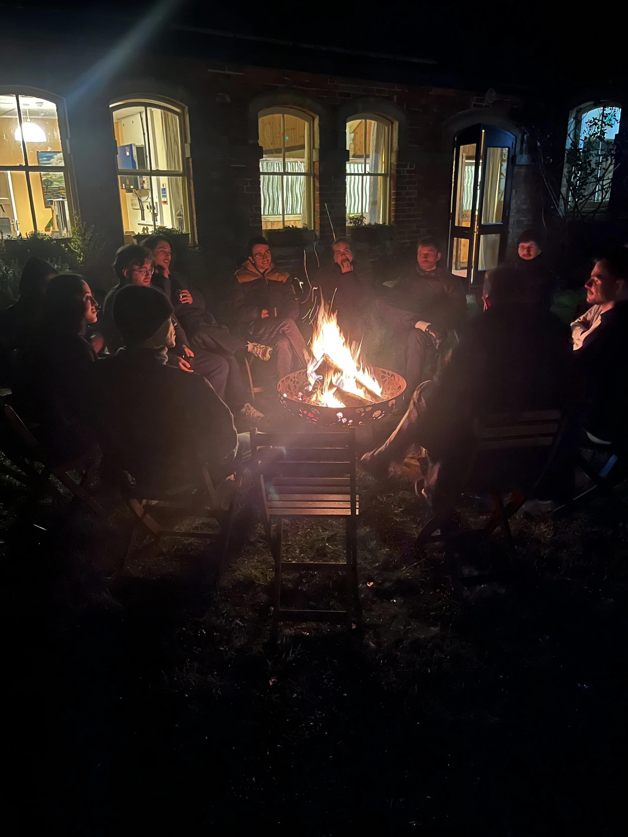 A group of people sitting around a campfire at night in a backyard, with a brick house and windows illuminated in the background.