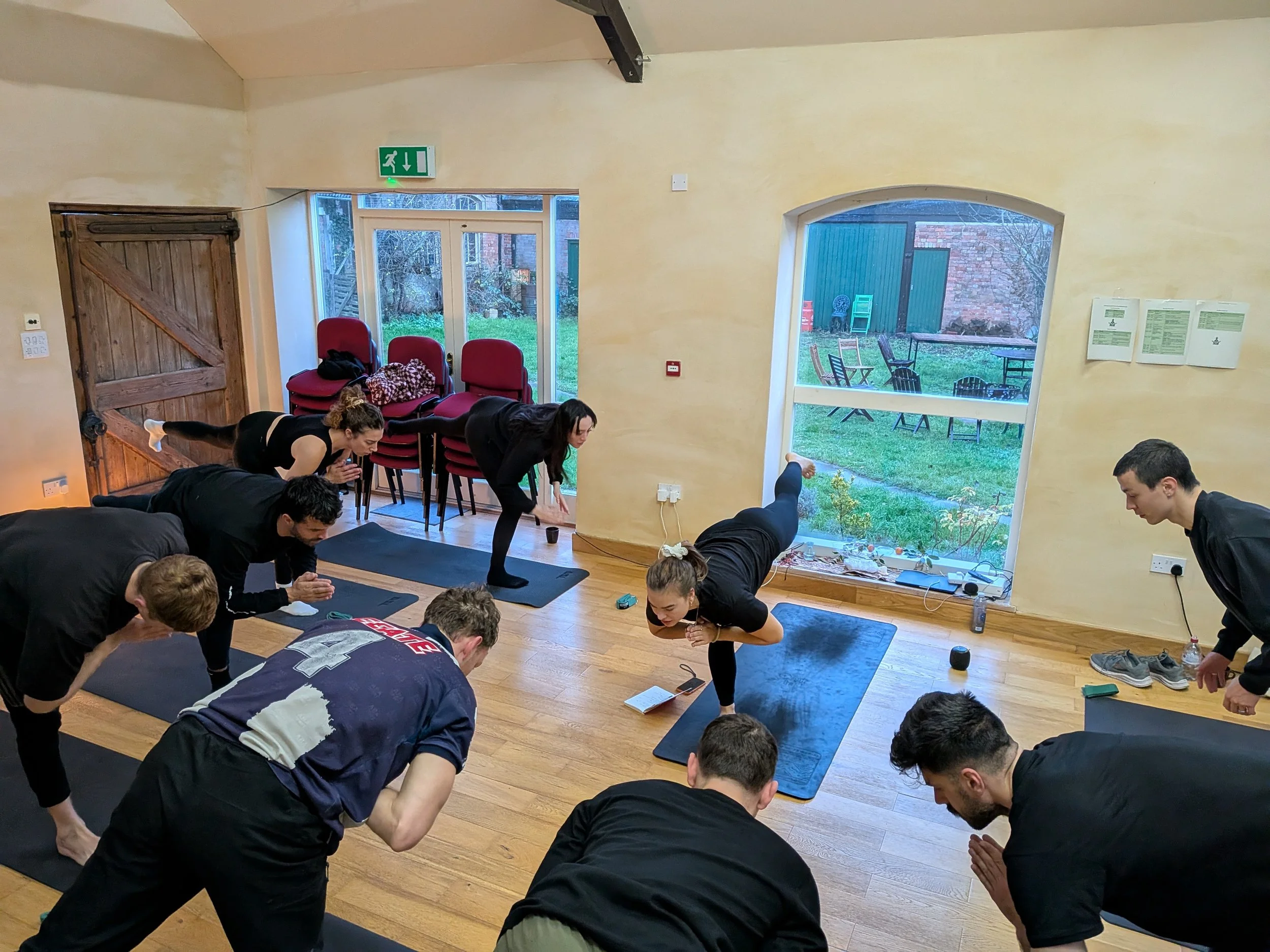 Indoor yoga or fitness class with multiple participants on mats practicing poses, in a room with large windows showing a garden outside, induction heating unit, chairs, and wall-mounted notices.