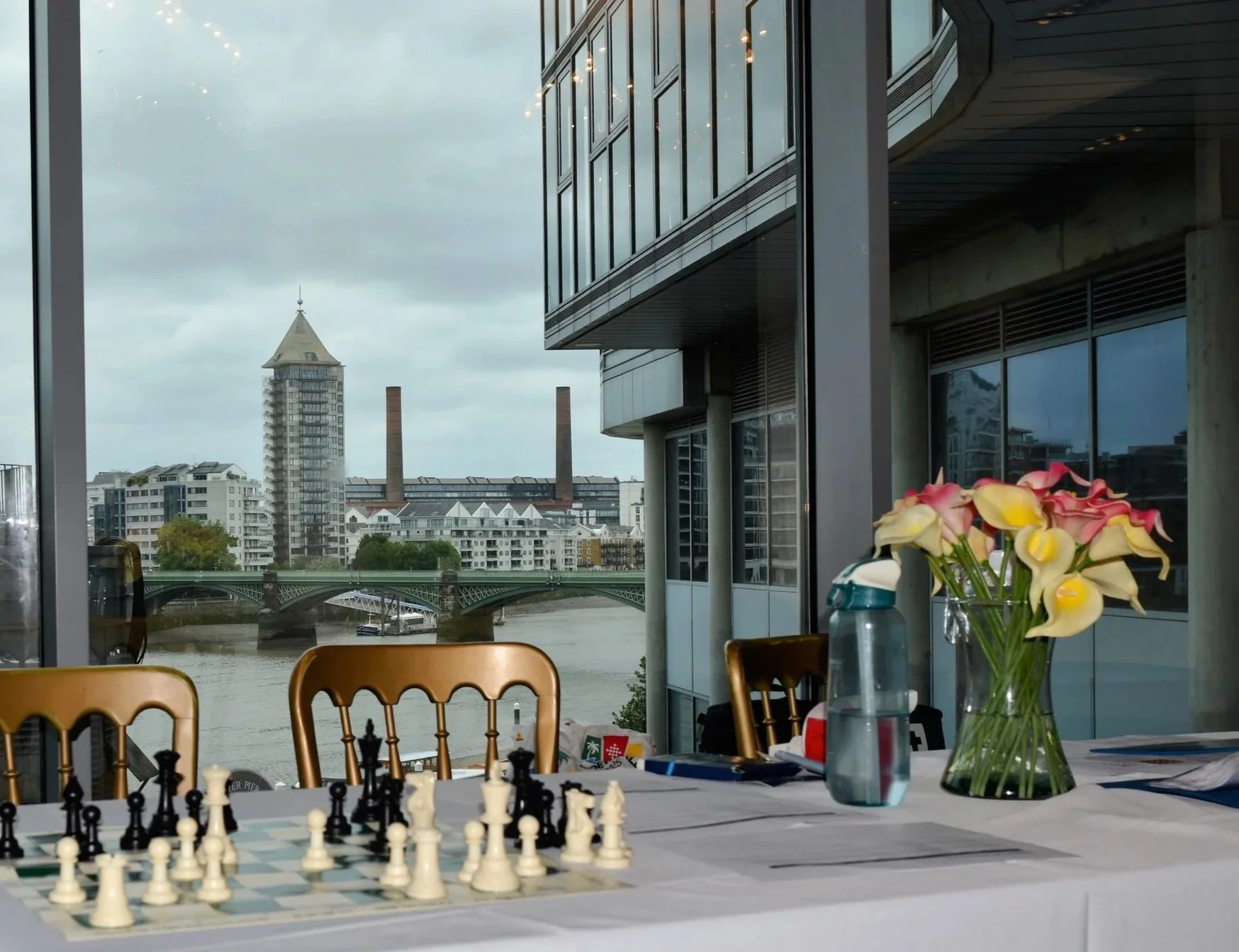Interior view of a modern apartment or office with a table set up for chess, a vase of calla lilies, and a water bottle. Large windows reveal a cityscape with high-rise buildings, a river, and an overcast sky.
