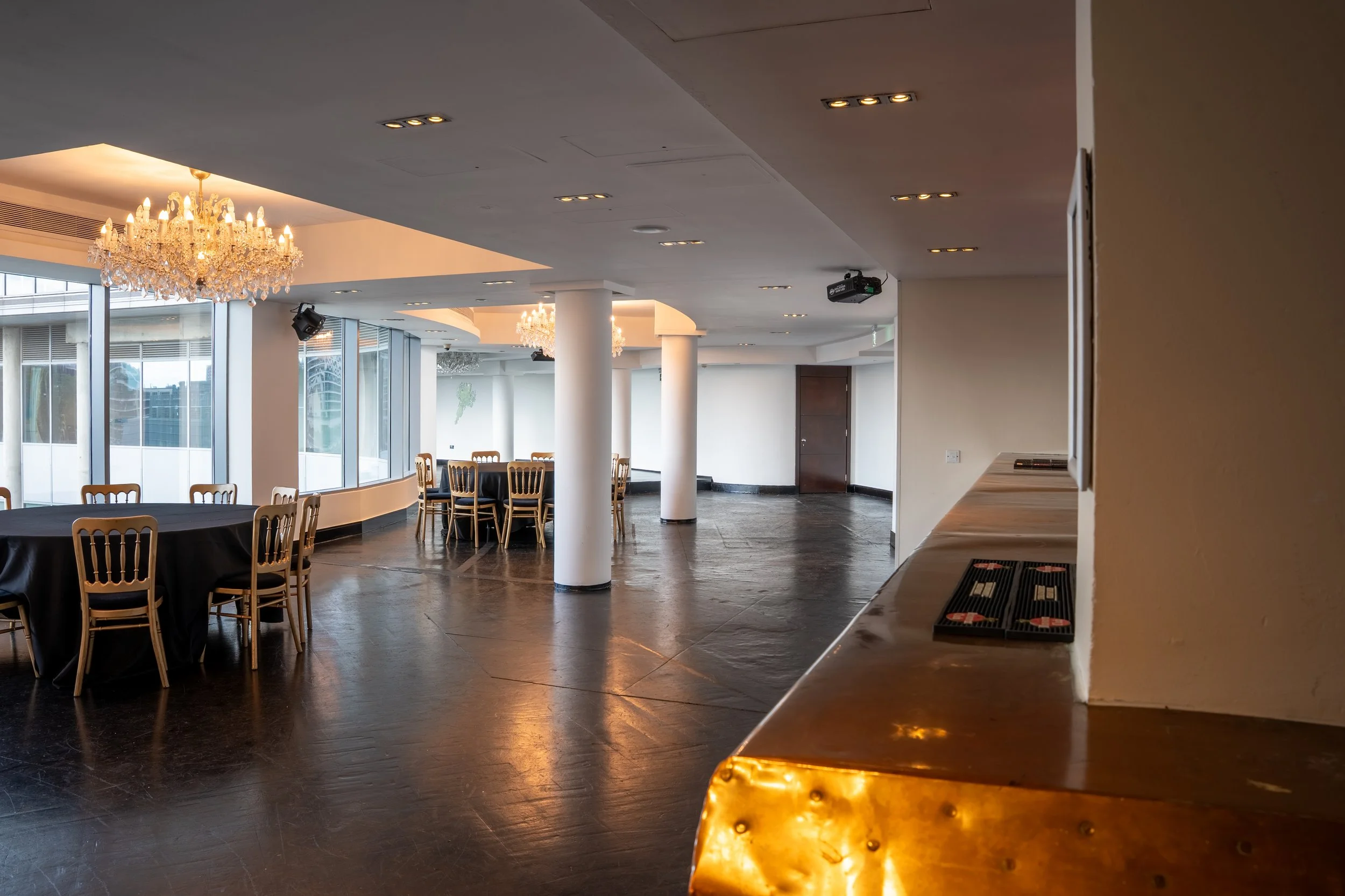 Spacious event room with large windows, chandeliers, and set tables with chairs, prepared for a gathering or banquet.