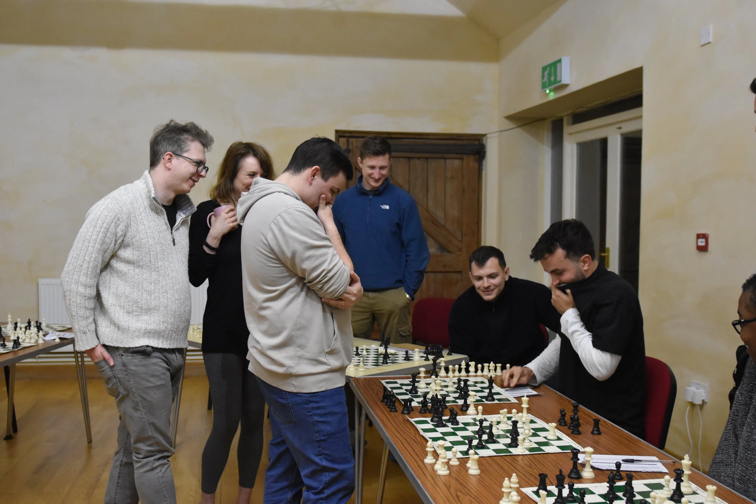 Group of people playing chess indoors, with some watching and others making moves, in a room with beige walls, wooden door, and emergency exit sign.