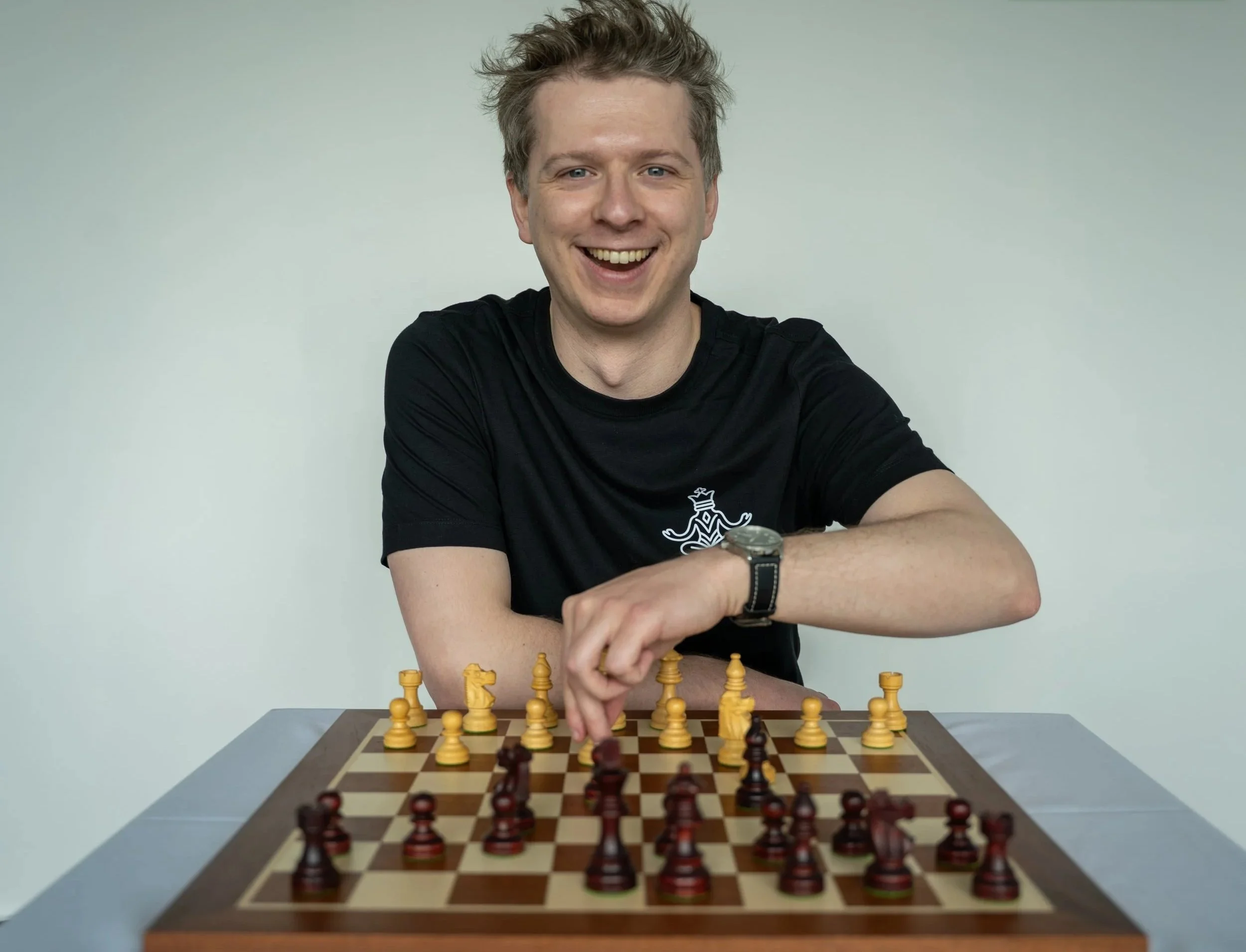 A man with short, messy hair smiling while playing chess at a table, wearing a black t-shirt with a small white logo, sitting in front of a chessboard with yellow and brown pieces, against a plain light-colored wall.