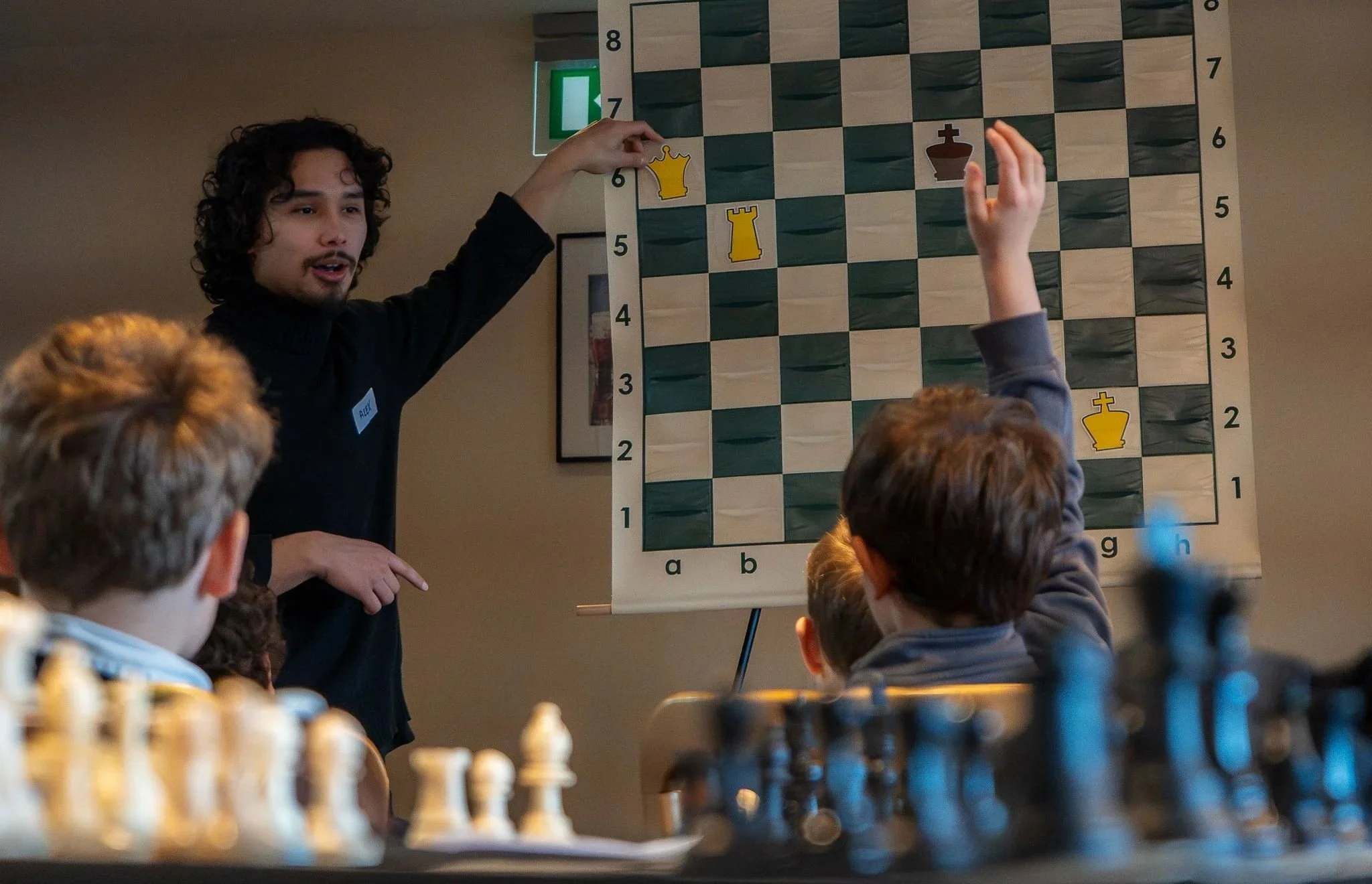 A person with curly hair and a black shirt explaining a chess game to children, with a chessboard on a poster behind them. Children are sitting at a table with chess pieces.