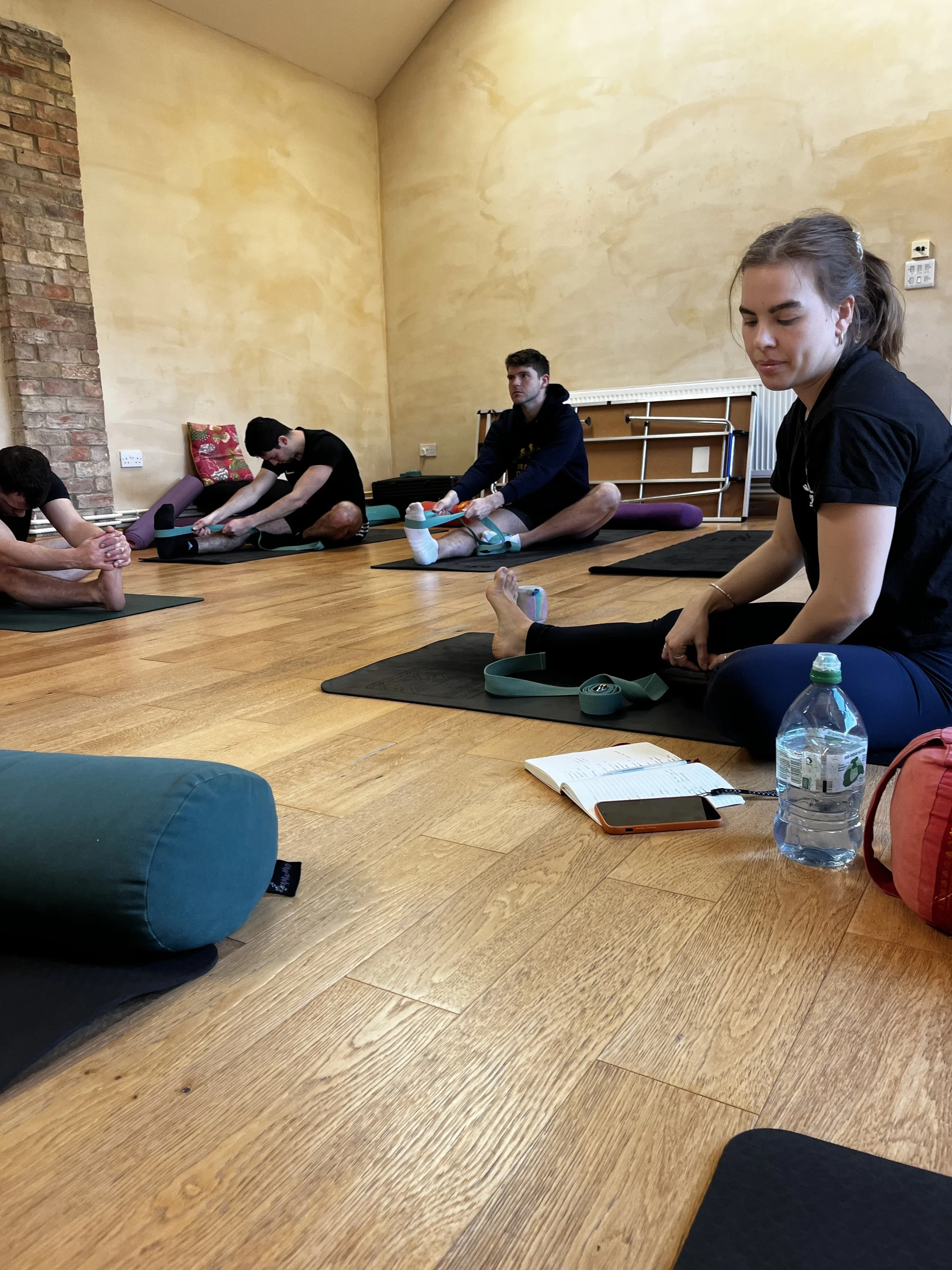 People participating in a seated yoga or meditation class in a room with wooden floors and yellow walls. Some are stretching or performing yoga poses, and there are yoga mats, a water bottle, a notebook, and a phone on the floor.