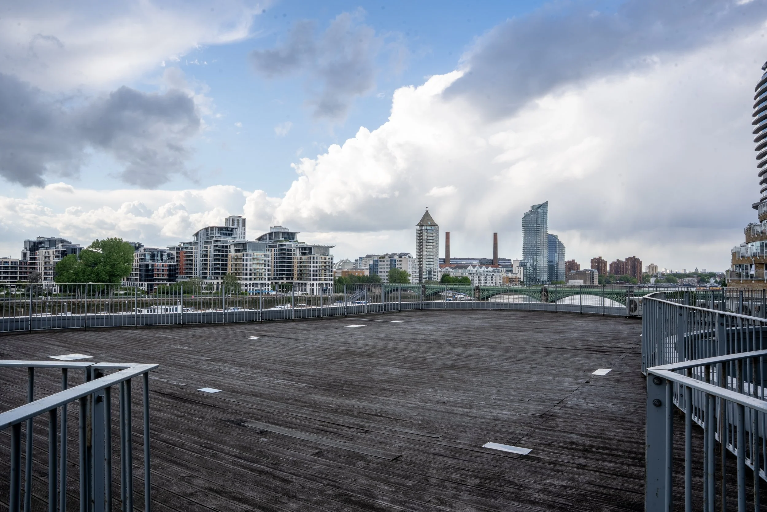 View of a city skyline with modern buildings, cloudy sky, and a rooftop deck with wooden flooring and metal railings.