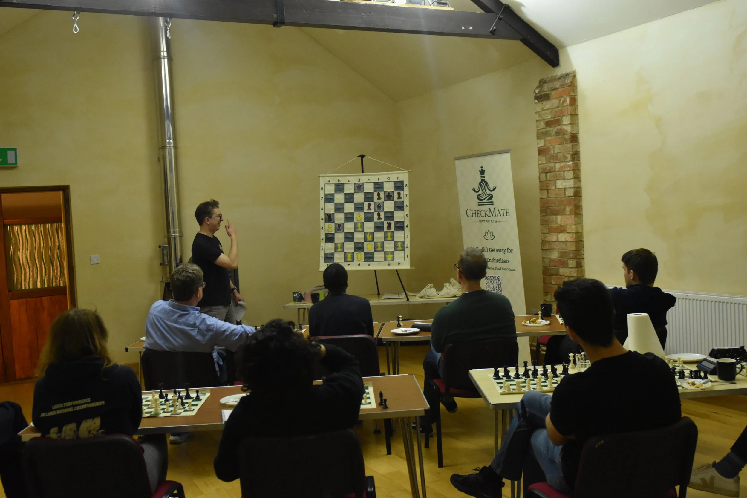 A group of people playing chess in a room with a yellow wall. A man is standing and gesturing in front of a large chessboard hanging on the wall, while others watch and play at tables. There is a banner with a chess logo and the word 'Checkmate Retre