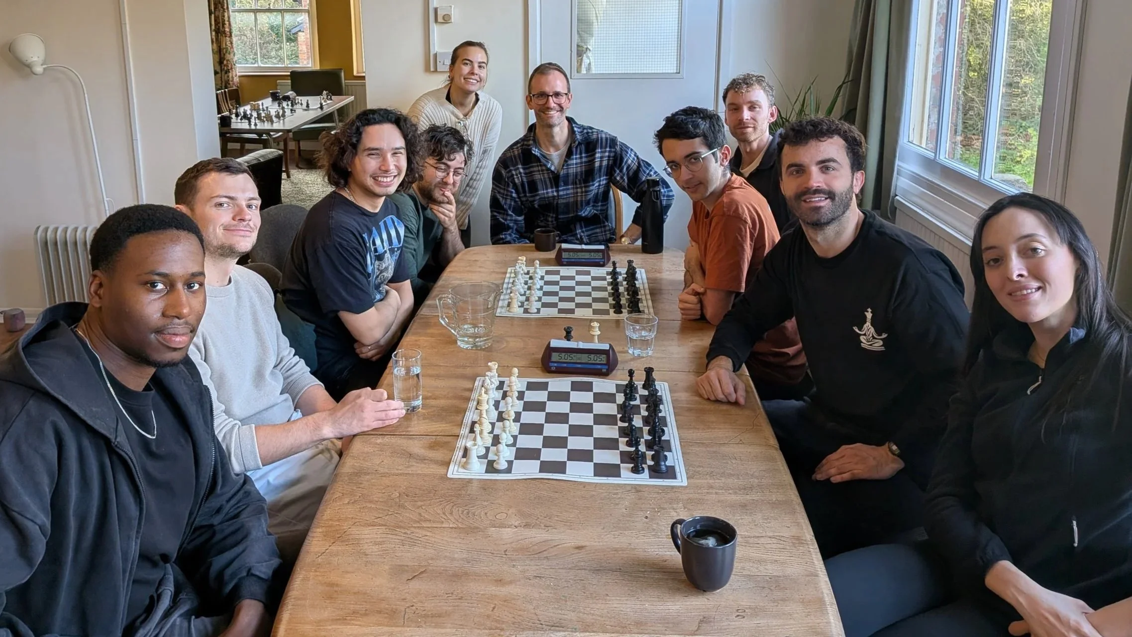 A group of ten people gathered around a table playing chess, smiling and looking at the camera in a well-lit room with large windows.