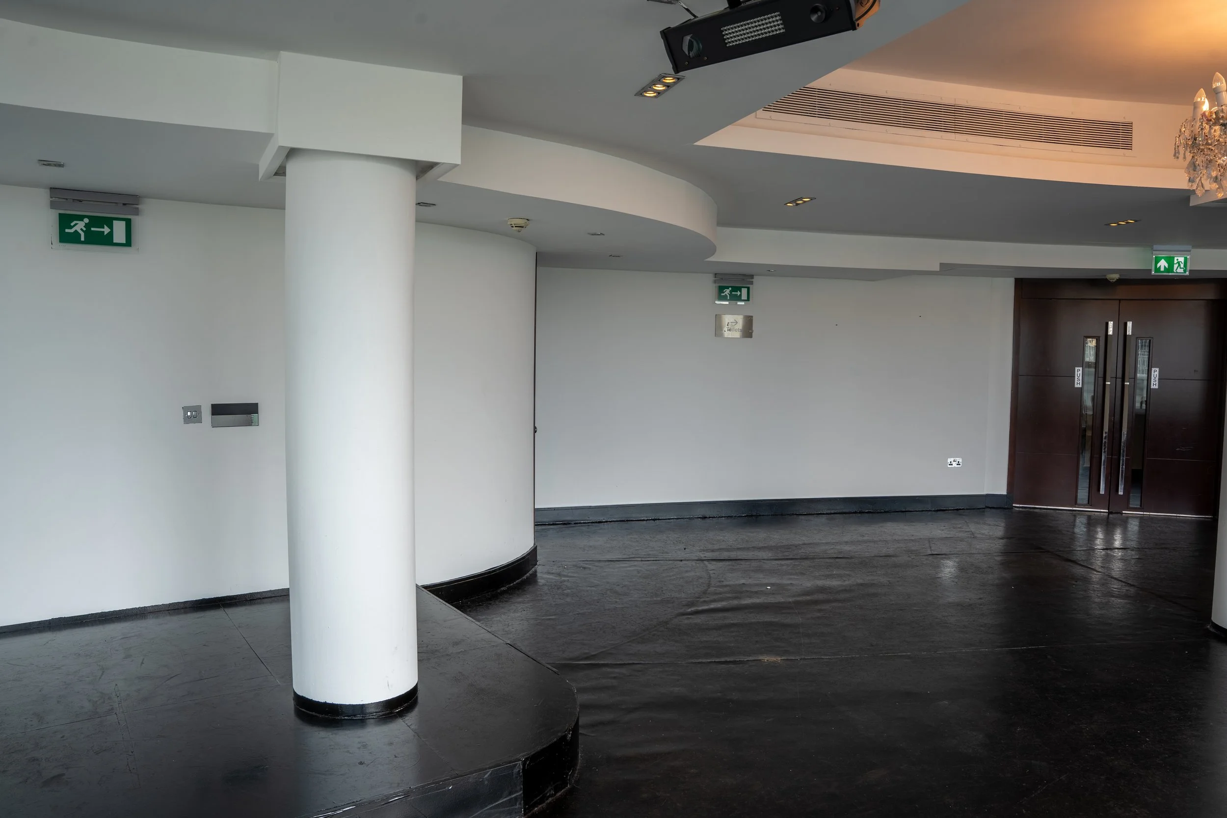 Interior of building with white walls, black stone flooring, a white round pillar, ceiling lights, emergency exit signs, and dark wooden doors.
