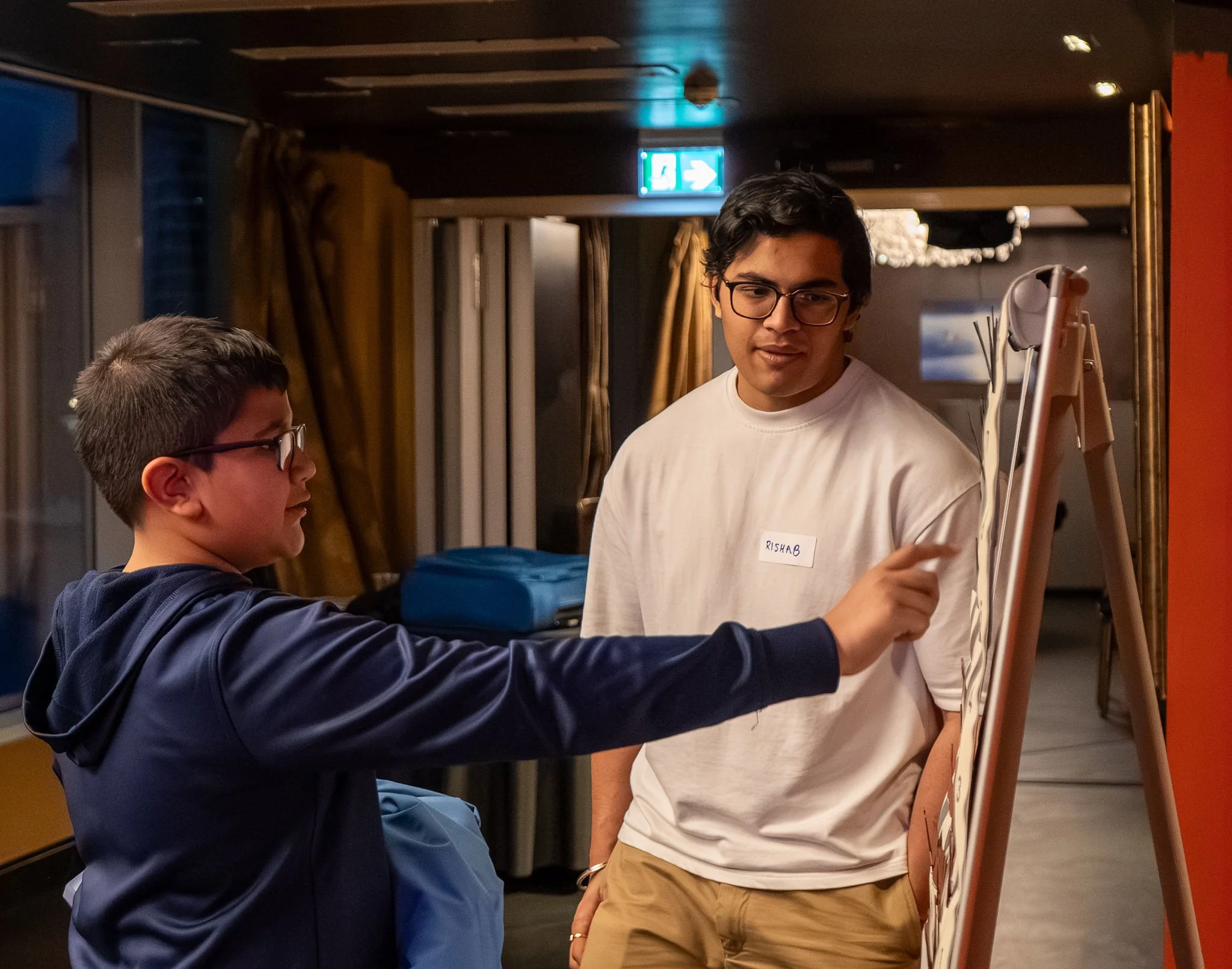Two young men, one with glasses and dark hair, and the other with short hair and glasses, standing near an easel or display board, engaged in conversation in a dimly lit room. The man with glasses is wearing a white shirt with a name tag that reads 'RISHAB'.