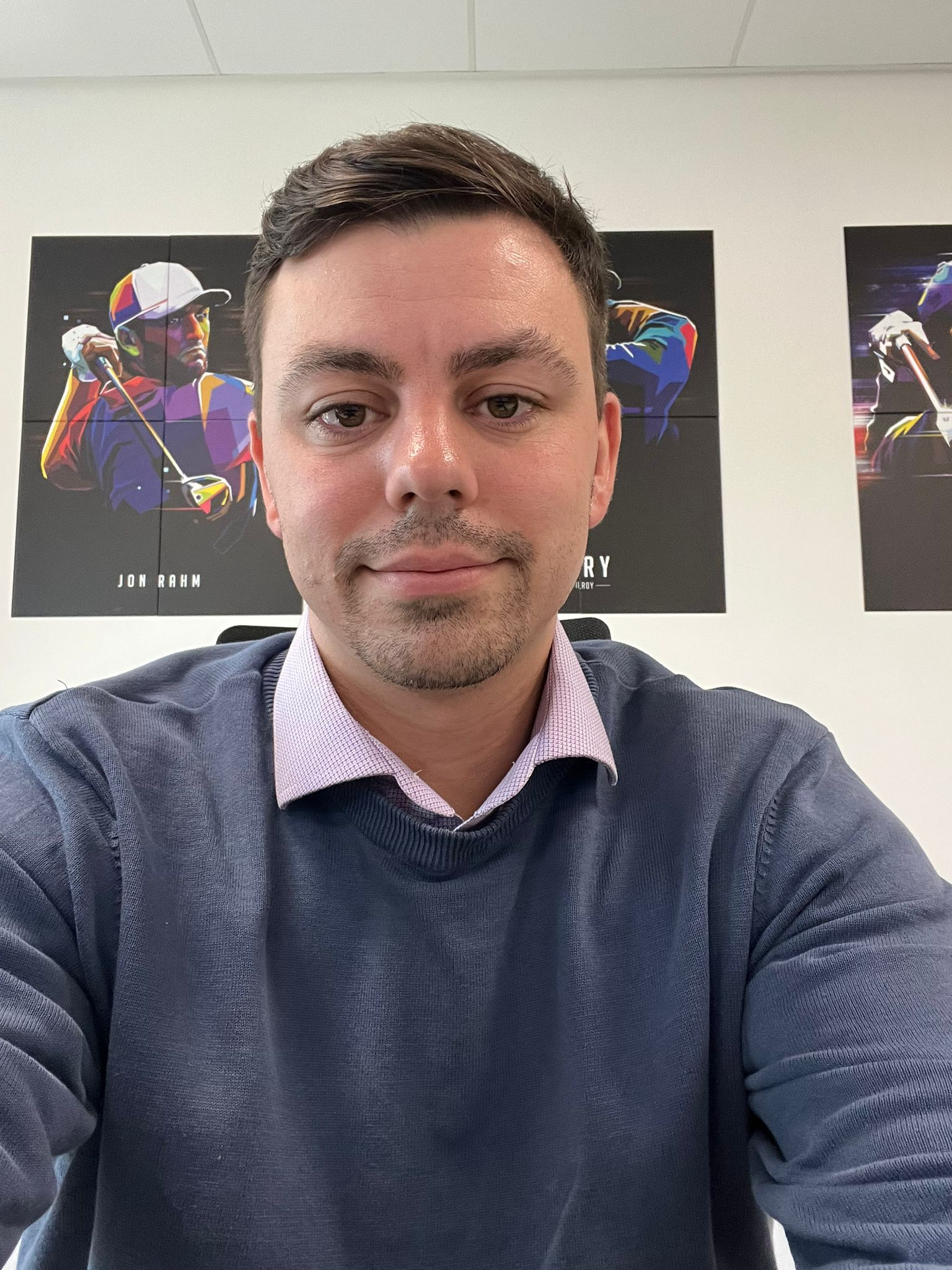A man with short brown hair and a slight beard taking a selfie in an office setting. Behind him are colorful posters of golfers. The man is wearing a dark navy sweater over a light purple collared shirt.