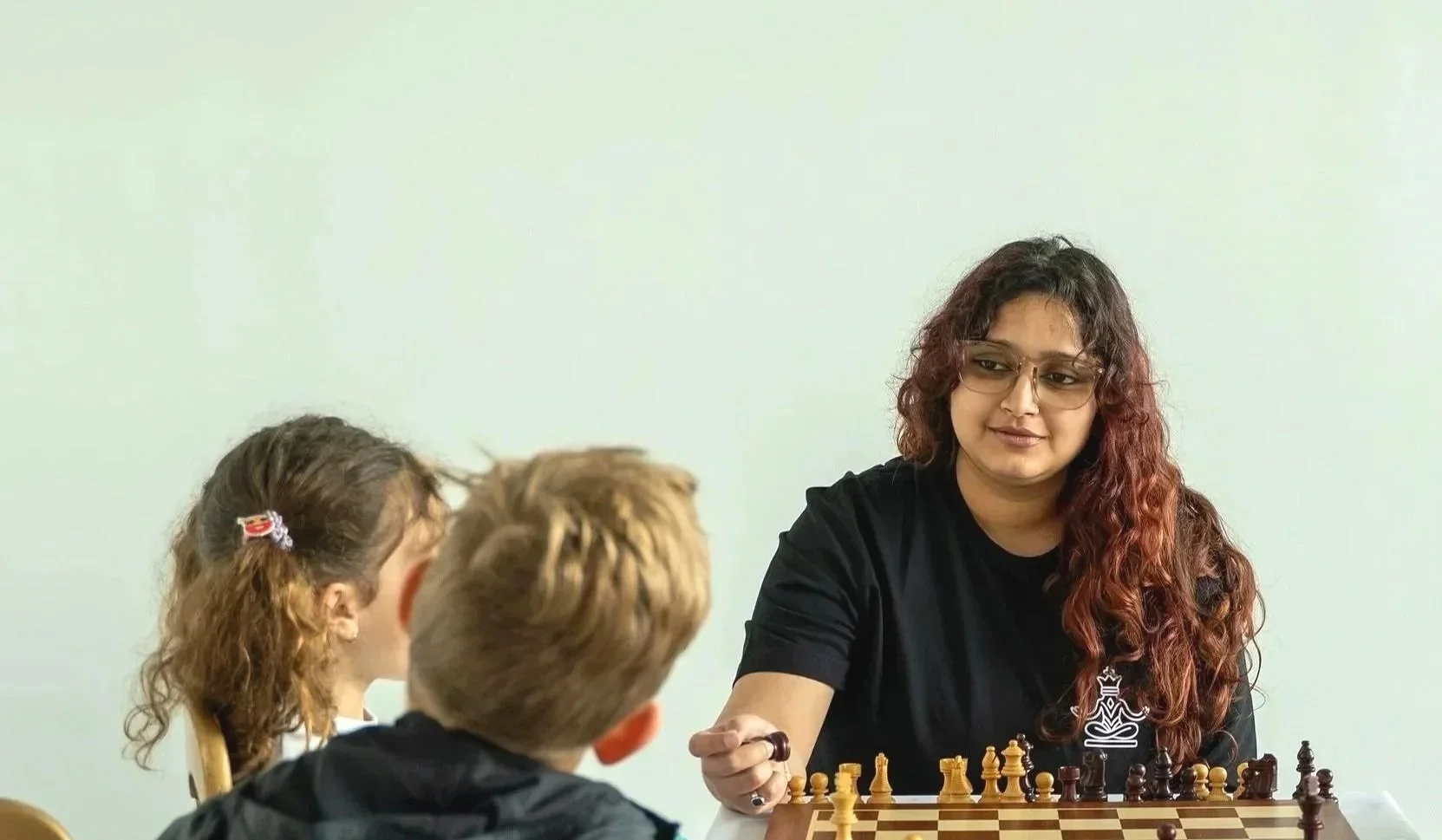A young woman in glasses with long, curly hair playing chess with three children at a table. The young woman is looking at the children, who are seated across from her, with their backs to the camera.