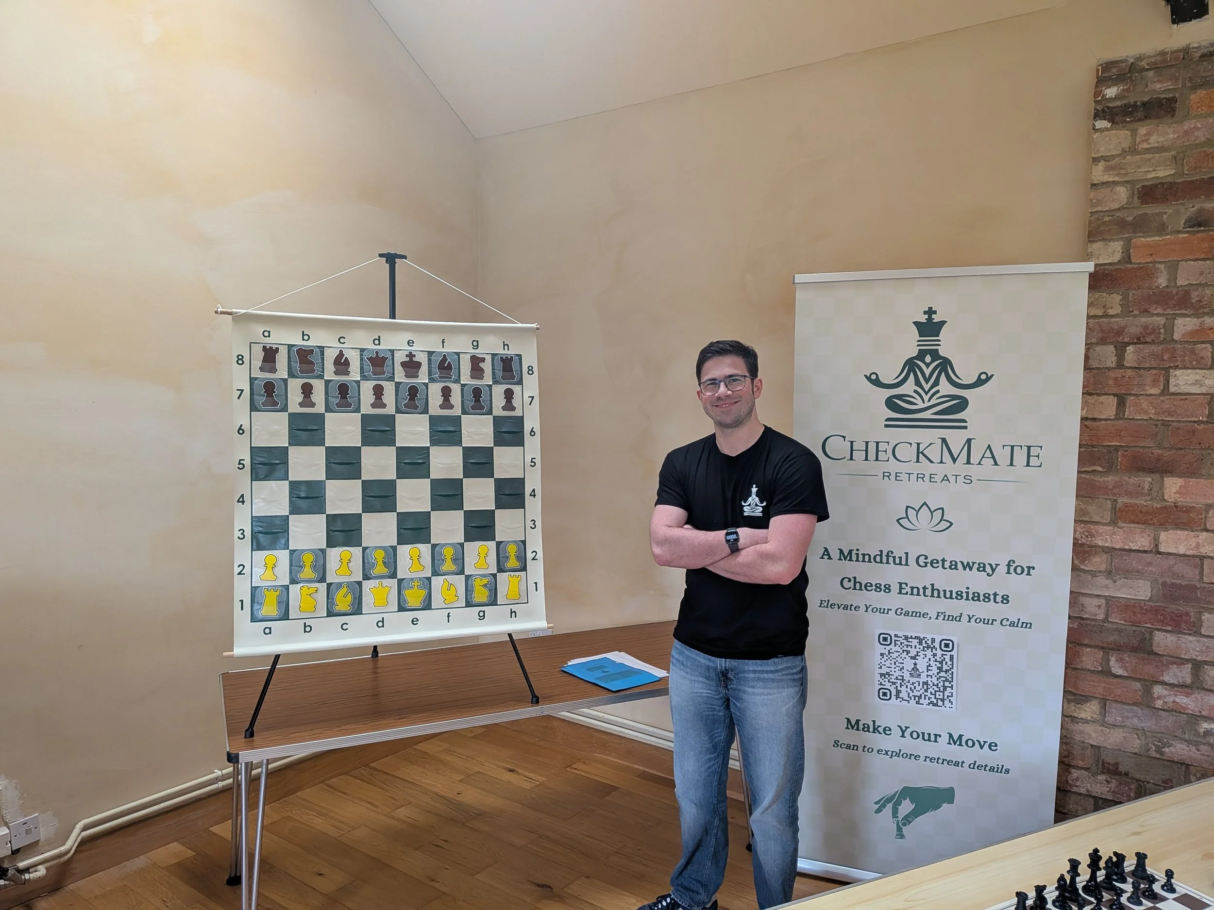 A man with glasses and dark hair, wearing a black t-shirt, stands with his arms crossed in front of a large chessboard and a promotional banner for CheckMate Retreats. The banner features a logo of a stylized figure with a crown and lotus flower, and