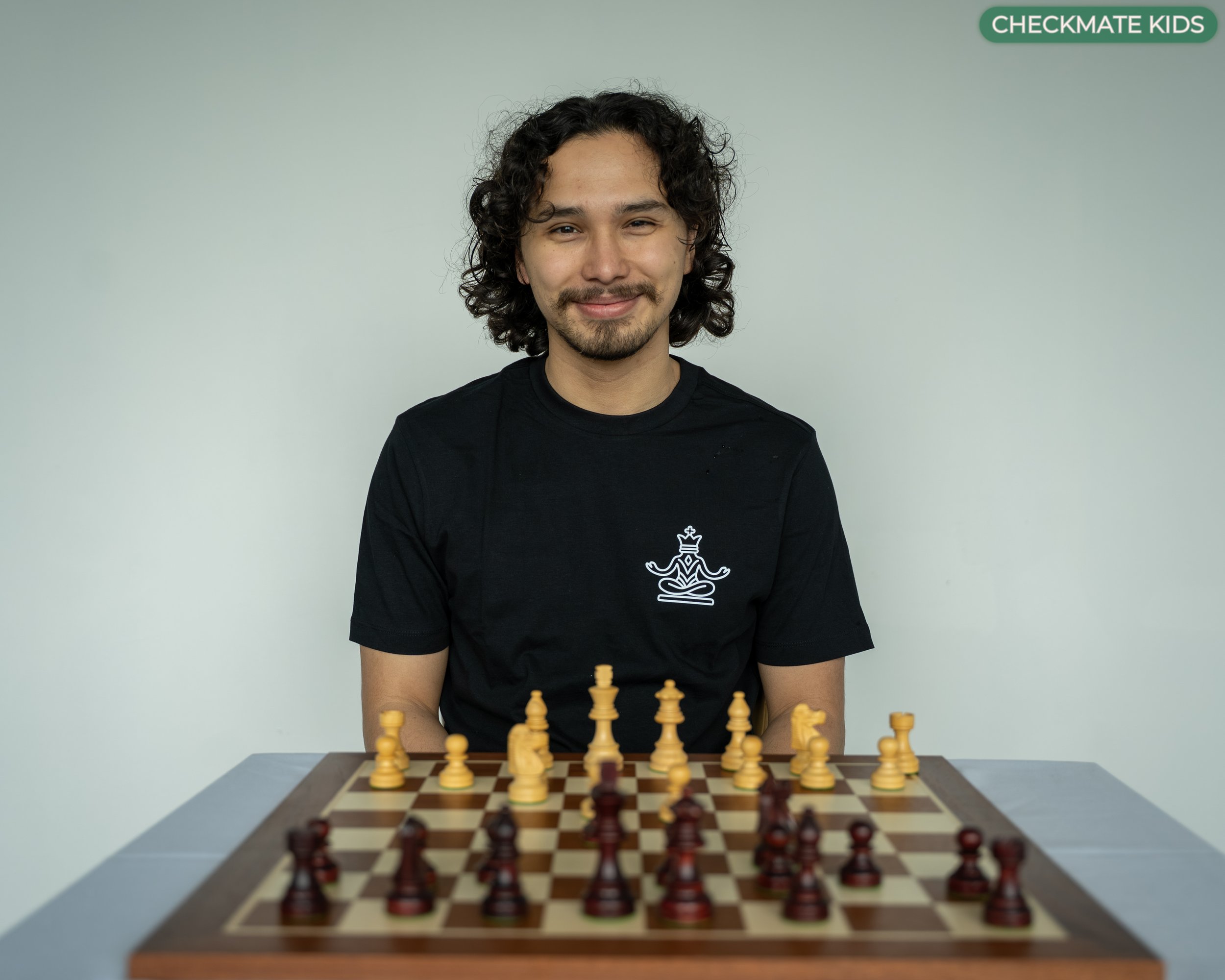 A young man with curly black hair, a mustache, and a goatee, wearing a black shirt with a chess piece symbol, sitting at a chessboard with chess pieces arranged for a game, in front of a plain light-colored wall.