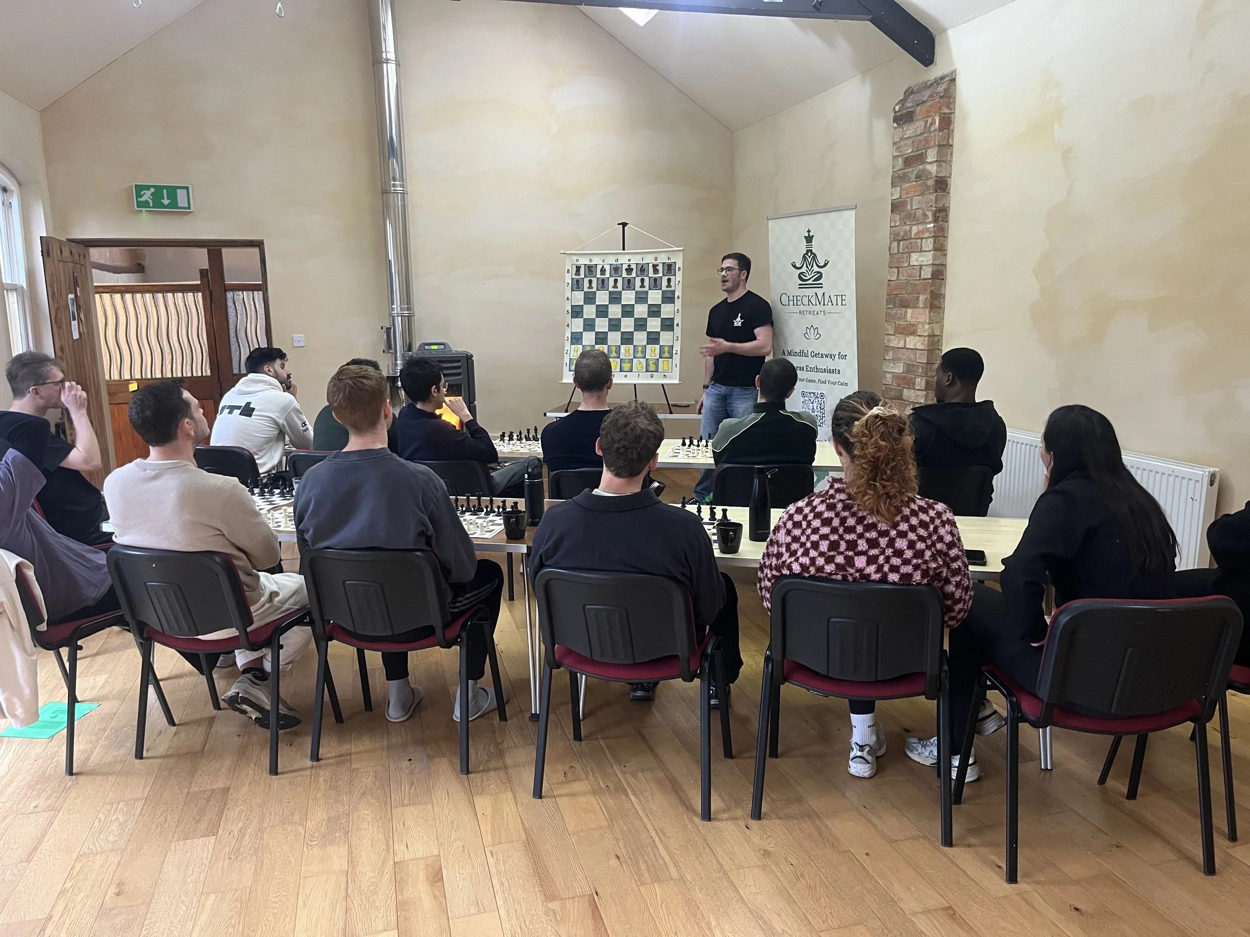 A group of people seated at tables in a room, listening to a man standing near a chessboard and a checkmate retreat banner, with some participants taking notes and others observing. The room has beige walls, wooden floors, and large windows.