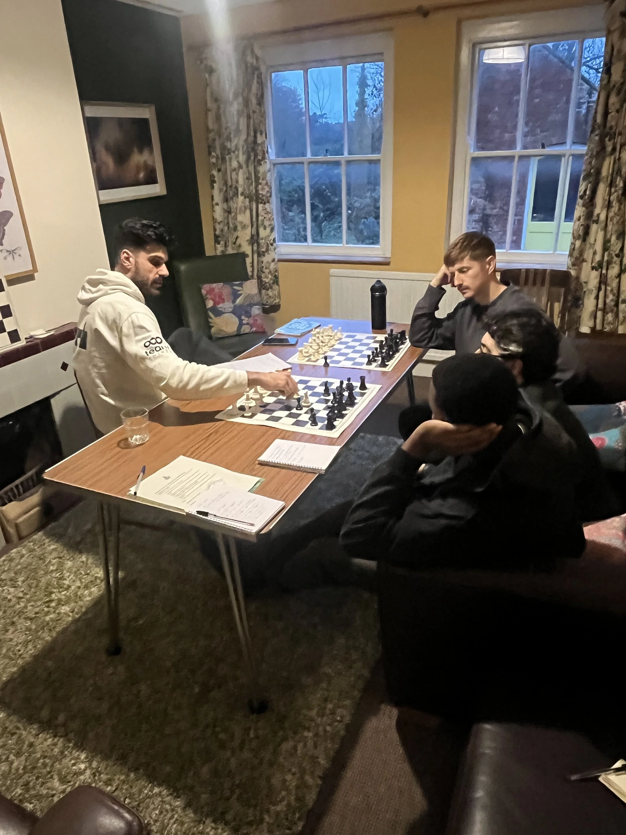 Two young men playing chess at a wooden dining table in a cozy room with yellow walls and large windows showing a dusk sky outside. There are papers, a water bottle, and a phone on the table, with some chairs and curtains nearby.