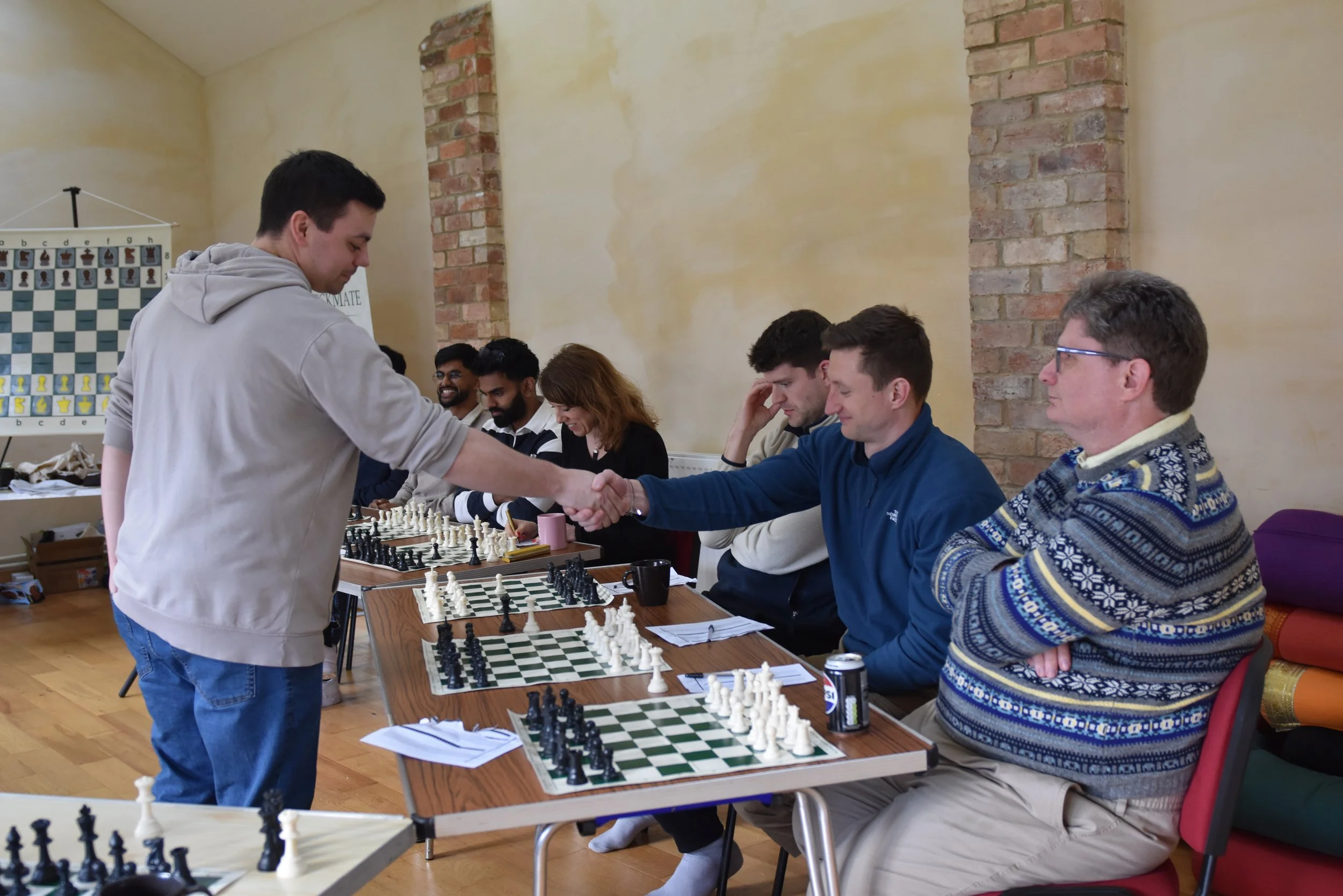 A group of people playing chess indoors, with one person in a gray hoodie shaking hands with another in a blue jacket, while others observe or think at the table.