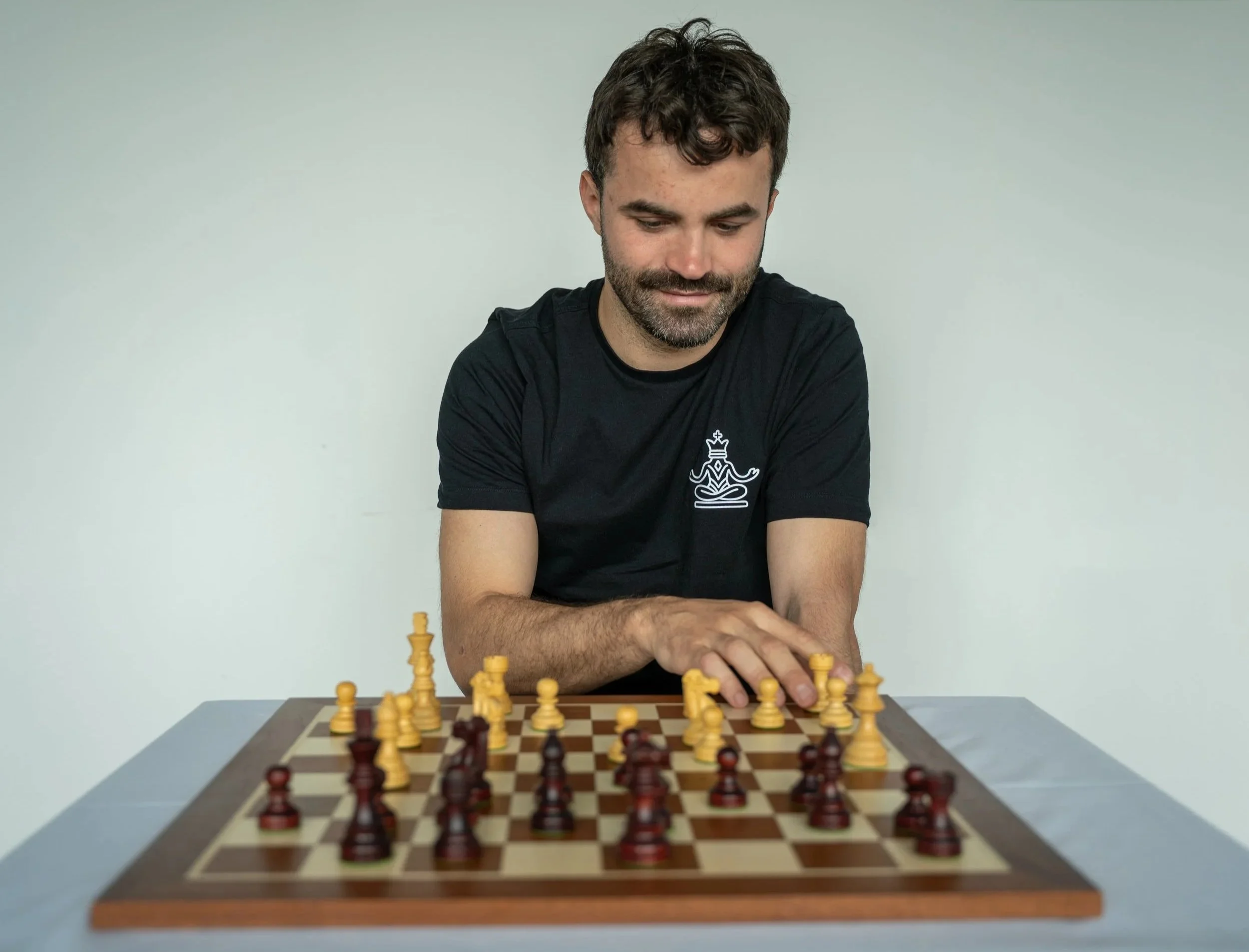 A man with dark hair and a beard is playing chess on a wooden chessboard. He is wearing a black shirt with a chess logo on it. The background is plain and white.