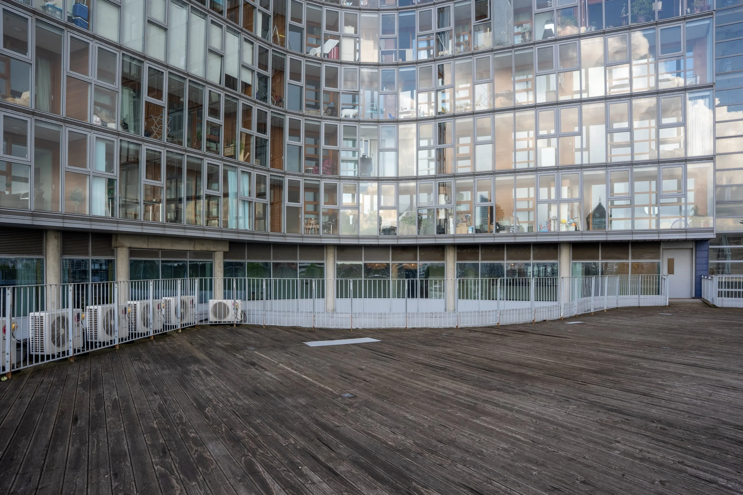 A modern glass building with curved facade and multiple floors, seen from a wooden outdoor terrace with a white metal railing and several air conditioning units.