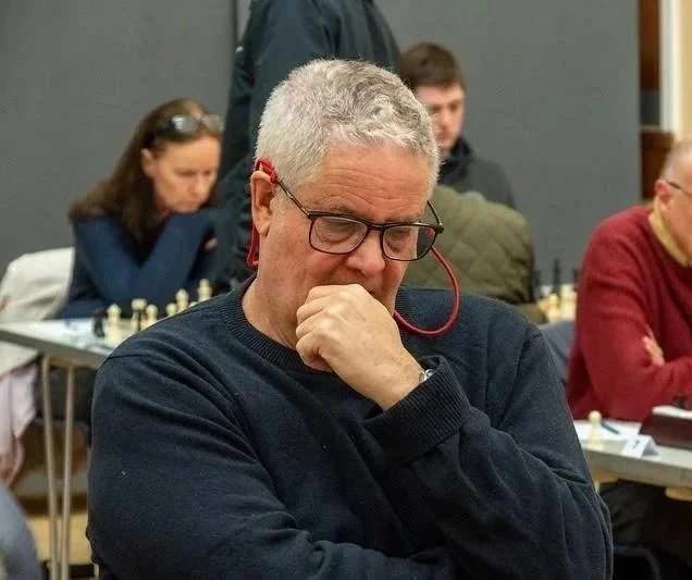 An older man with gray hair, glasses, and headphones is deep in thought during a chess game in a competitive setting, with other players visible in the background.