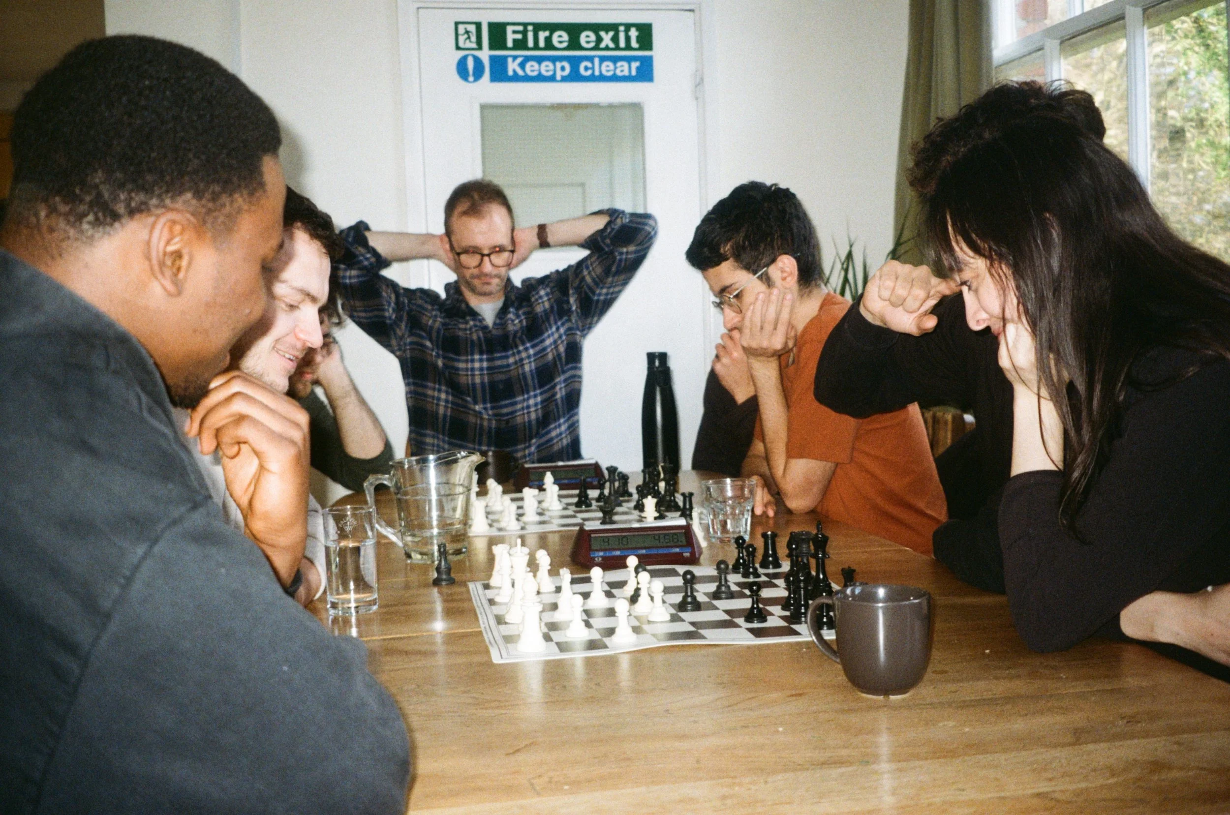 A group of people playing chess at a table in a room, with two of them appearing deep in thought and others watching the game. There is a chess clock and glasses of water on the table, and a window with greenery outside.