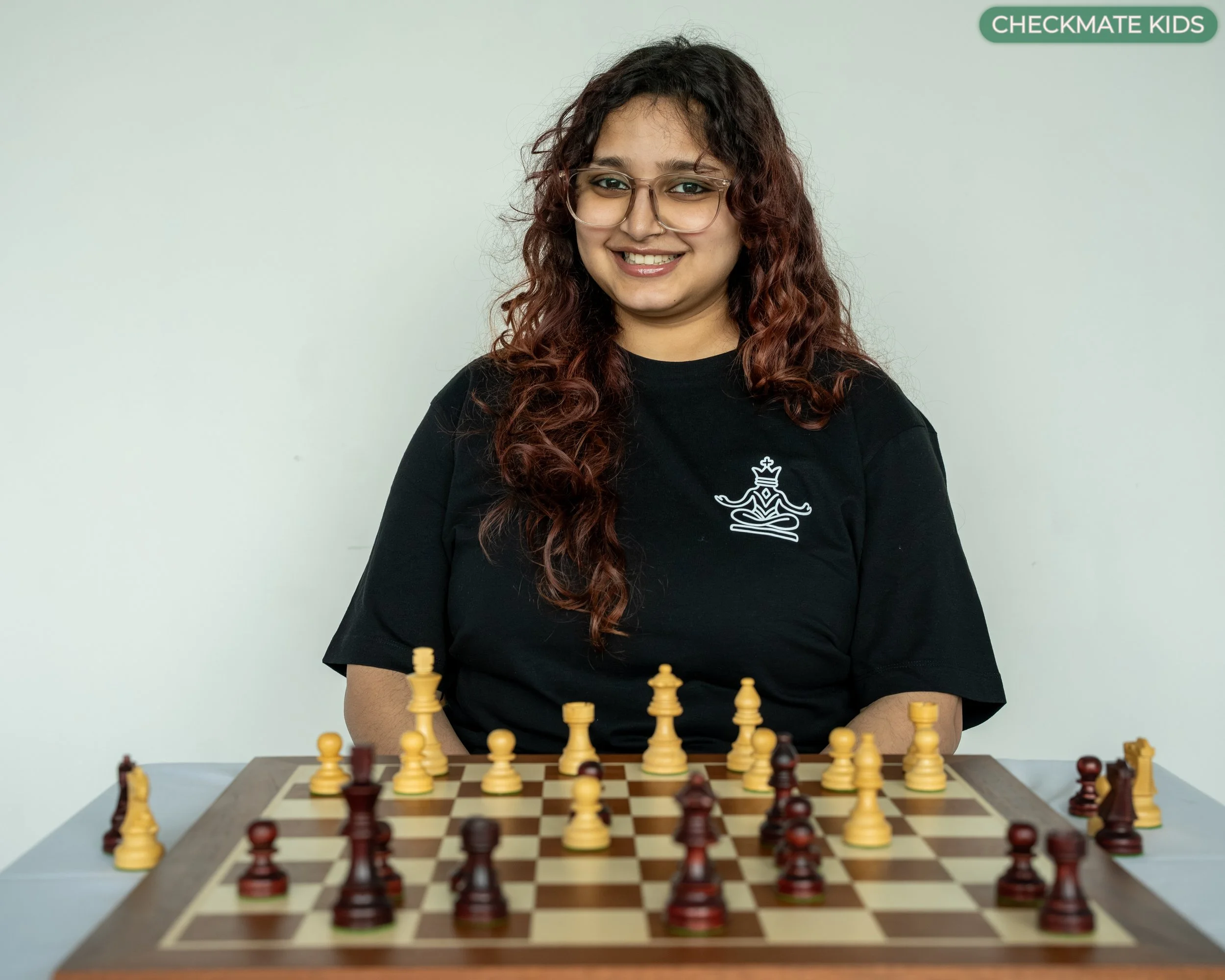 A young woman with long curly hair, glasses, and a black shirt with a chess logo, playing chess.
