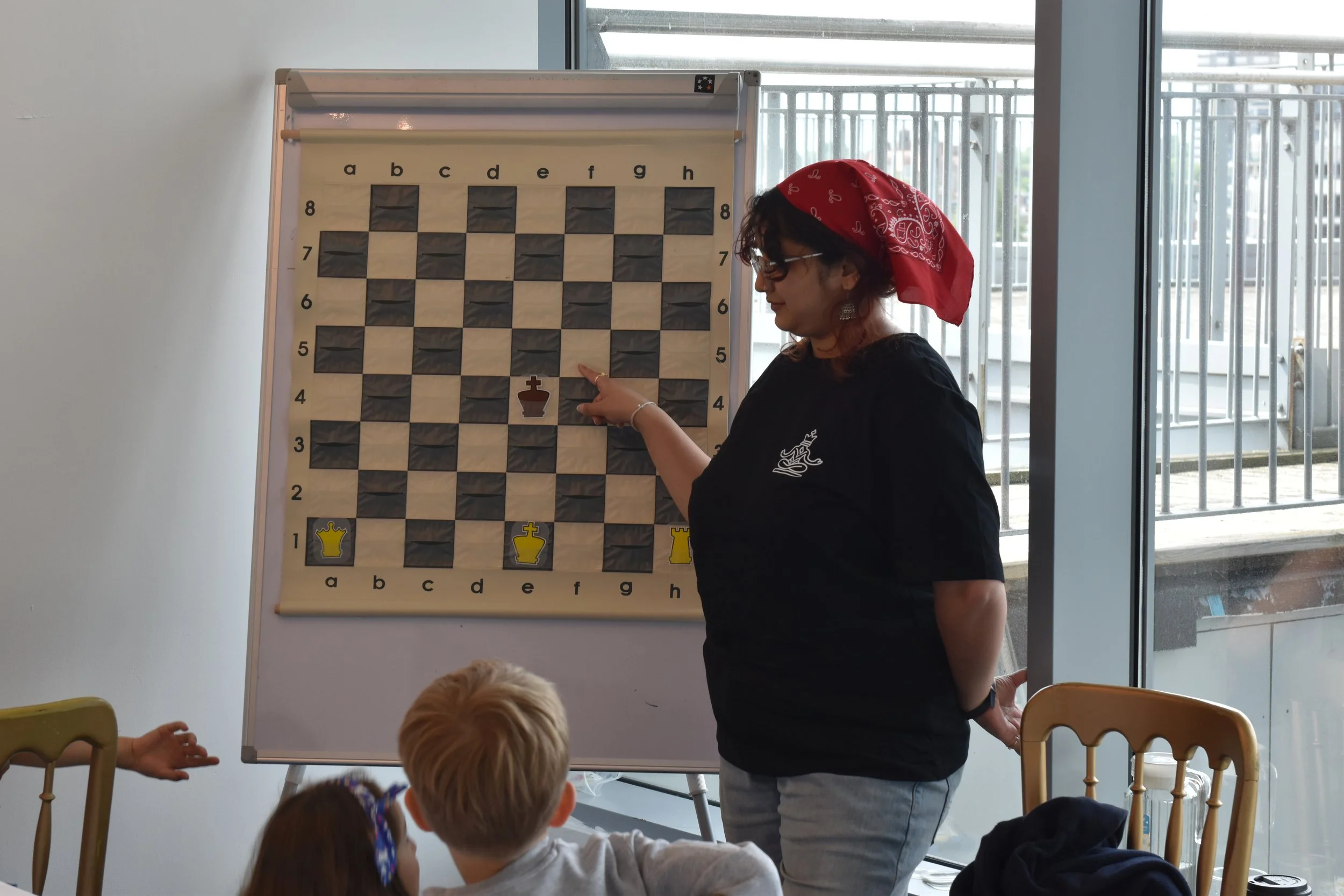 A woman in a red bandana and glasses is pointing at a chessboard on a whiteboard, with children seated at tables watching. The chessboard has an ongoing game.