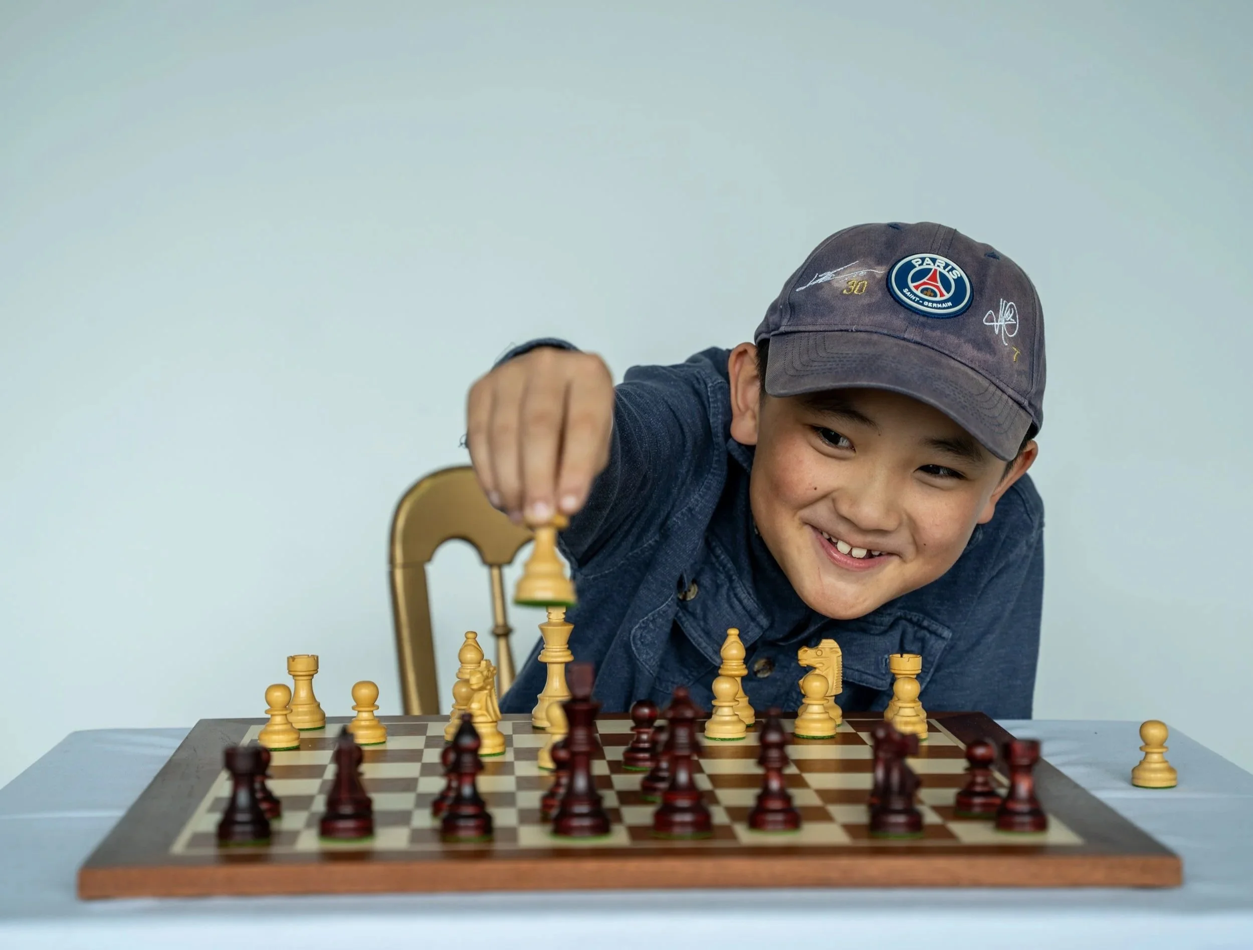 A young person playing chess at a table with a chessboard and pieces, wearing a gray cap with patches and symbols, smiling, against a plain light blue background.