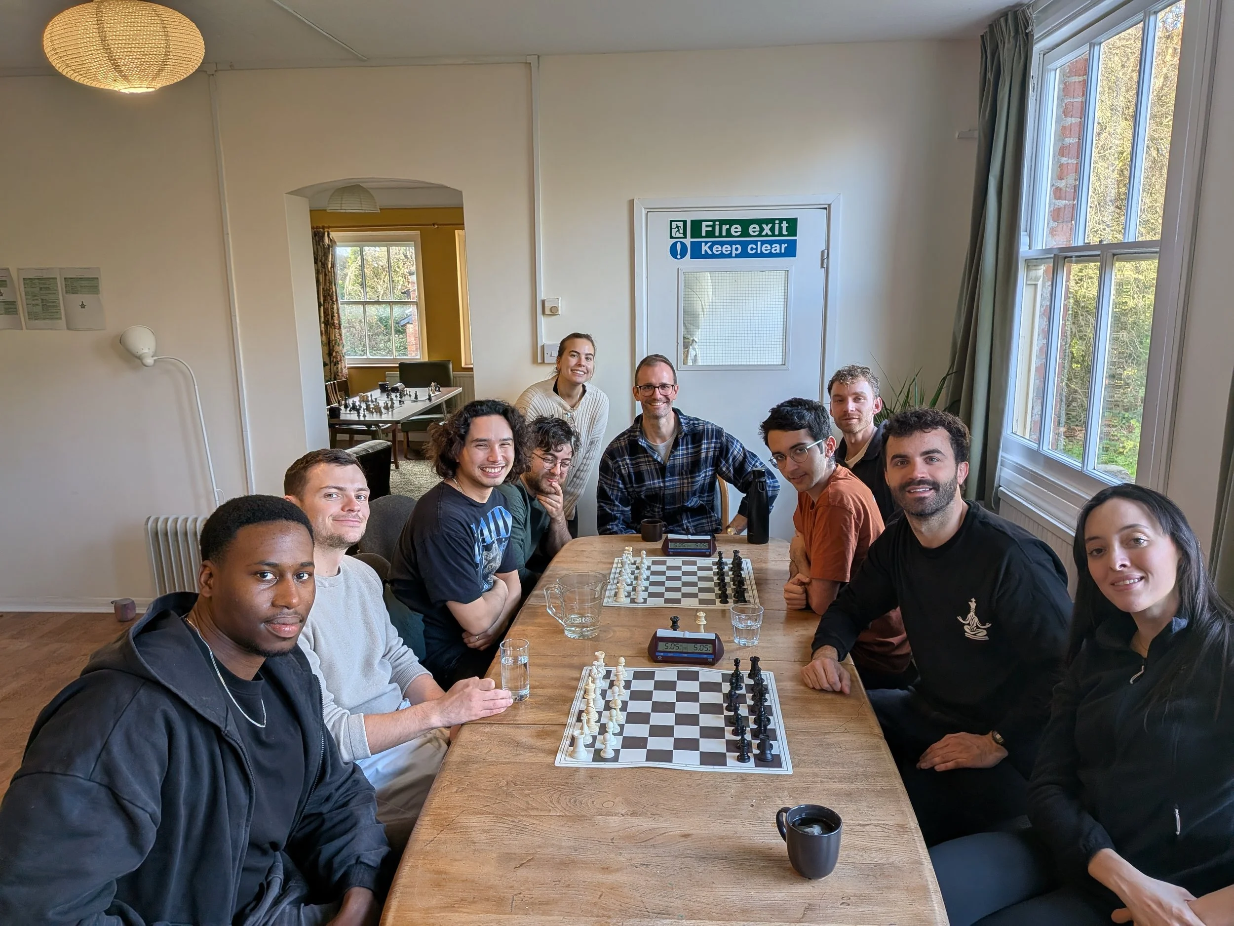 Group of people sitting around a table playing chess inside a room with large windows, sunlight, and a chess clock on the table.