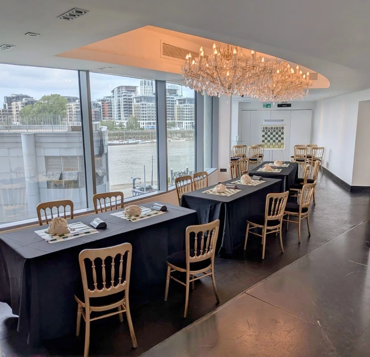A restaurant or event space with large windows overlooking a river and modern buildings. There is a chandelier hanging from the ceiling, and a long table with black tablecloths, chairs, and neatly folded napkins.
