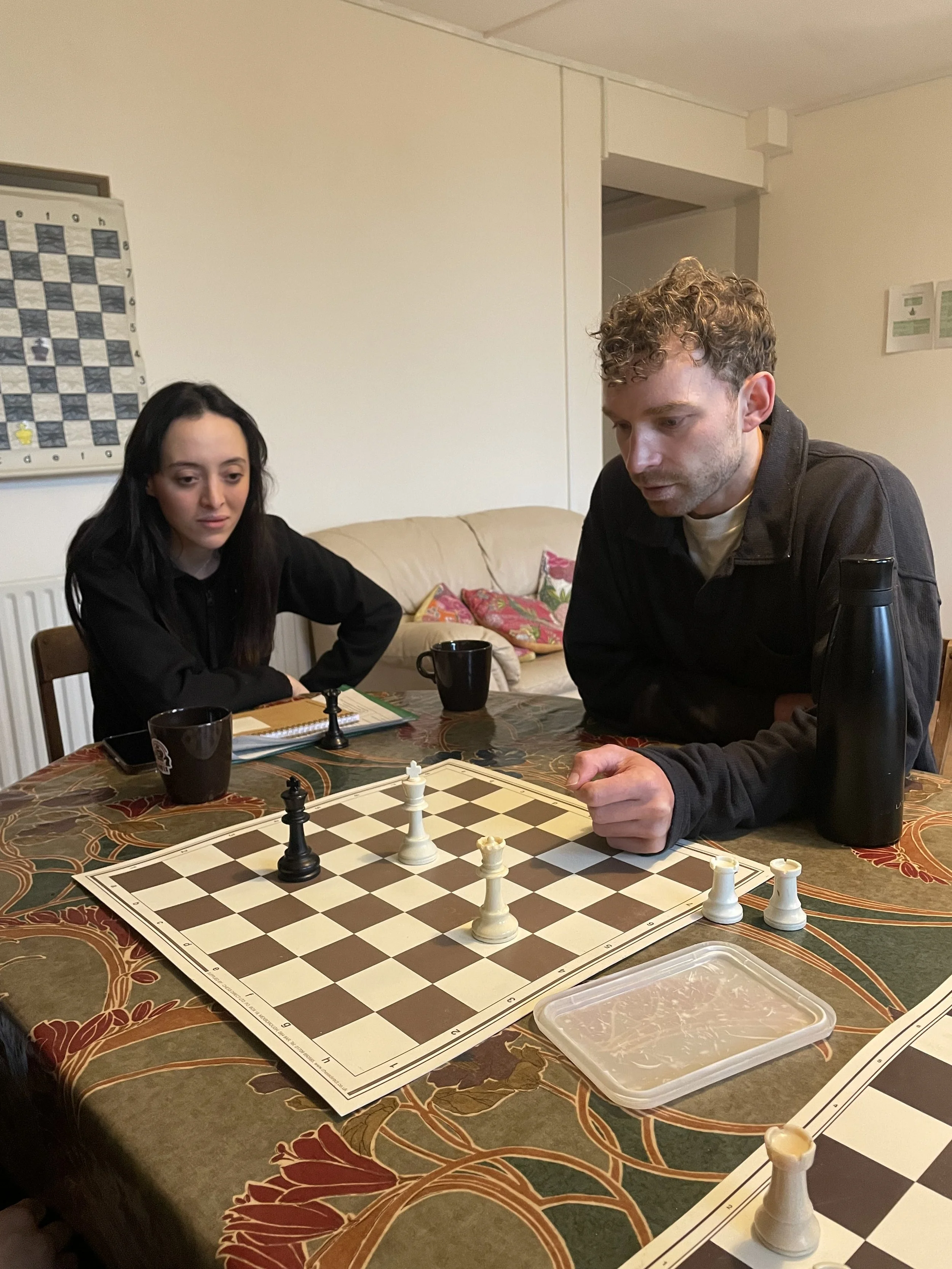 A man and woman are playing chess at a table covered with a floral tablecloth. The man is moving a white pawn, while the woman observes. The chessboard is in the center, and there are two black coffee mugs on the table. A thermos or water bottle is a