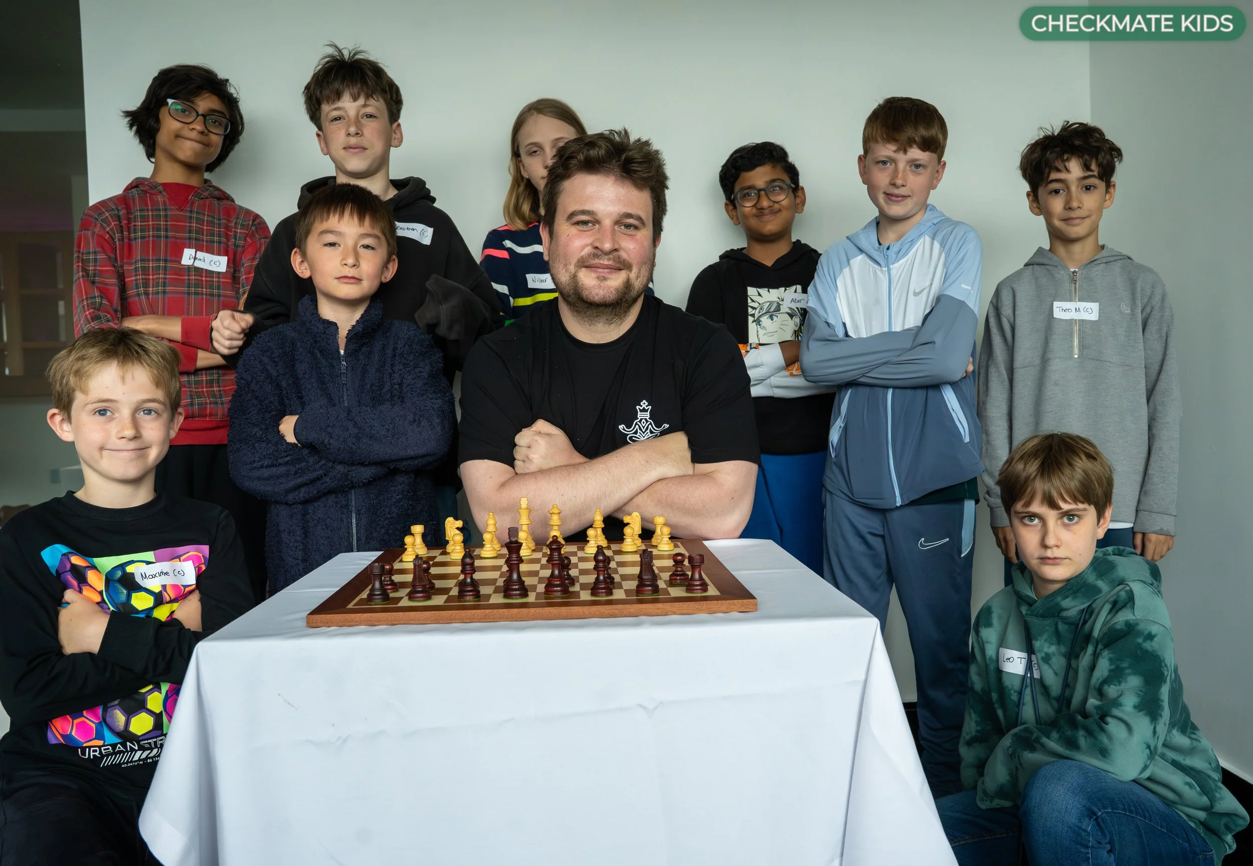 A group of children and an adult man posing behind a chessboard during a chess event at a school or community center, with all individuals wearing casual clothes and some children wearing name tags.