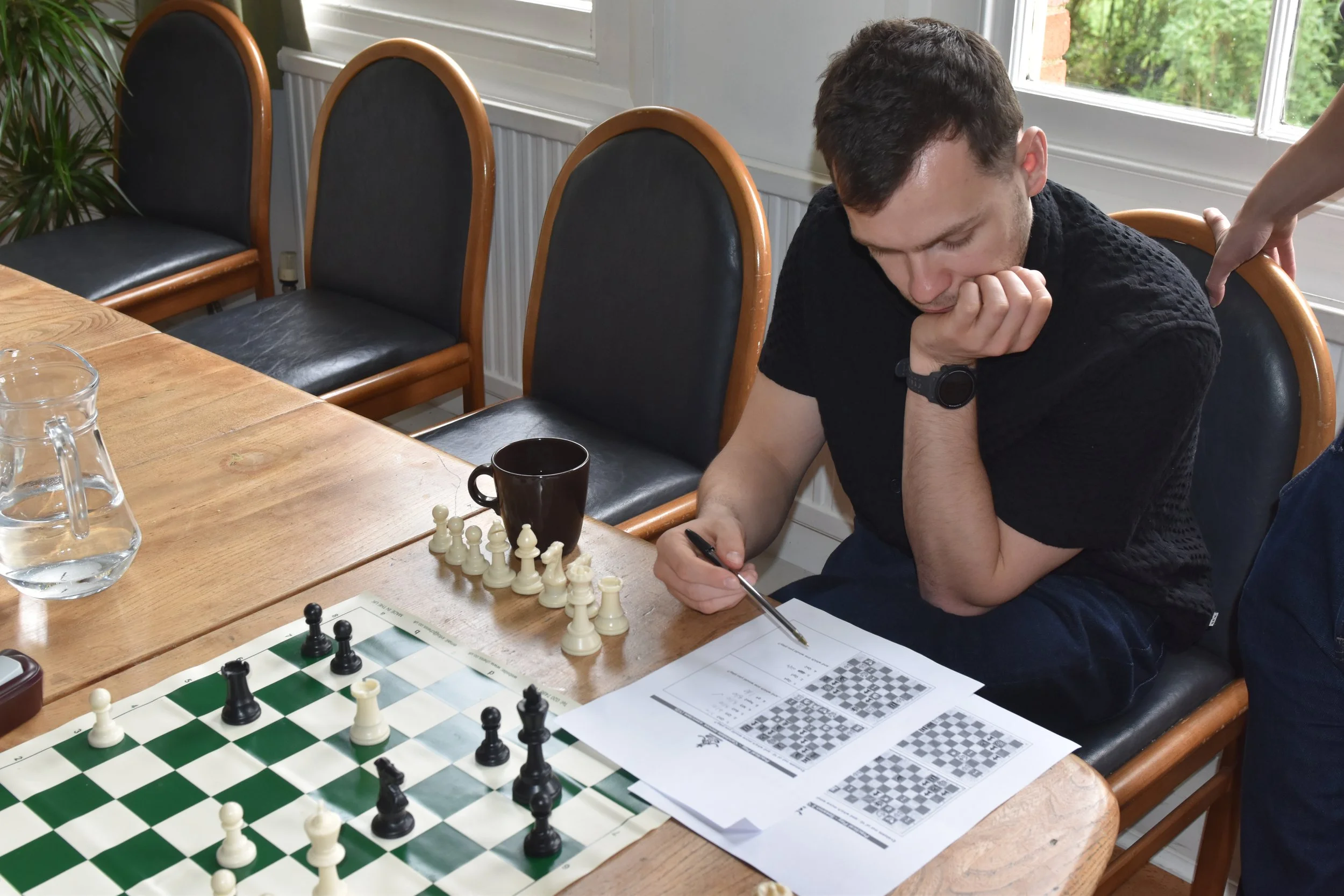 A young man sits at a wooden table, focusing on a chess game, with a pen in his hand and papers nearby, in a sunlit room with chairs and a window in the background.