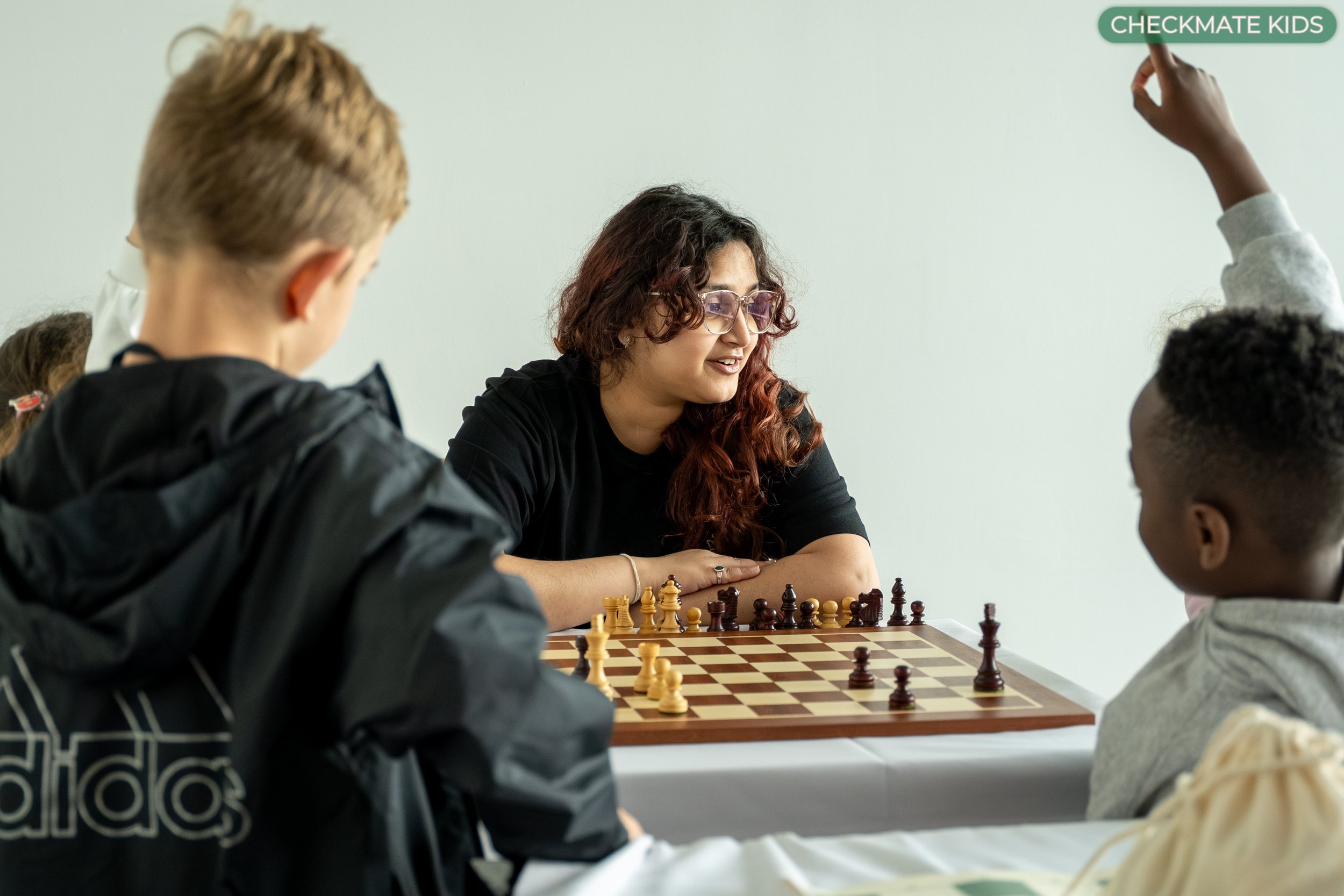 Young woman with curly hair and glasses playing chess with a group of children in a classroom.