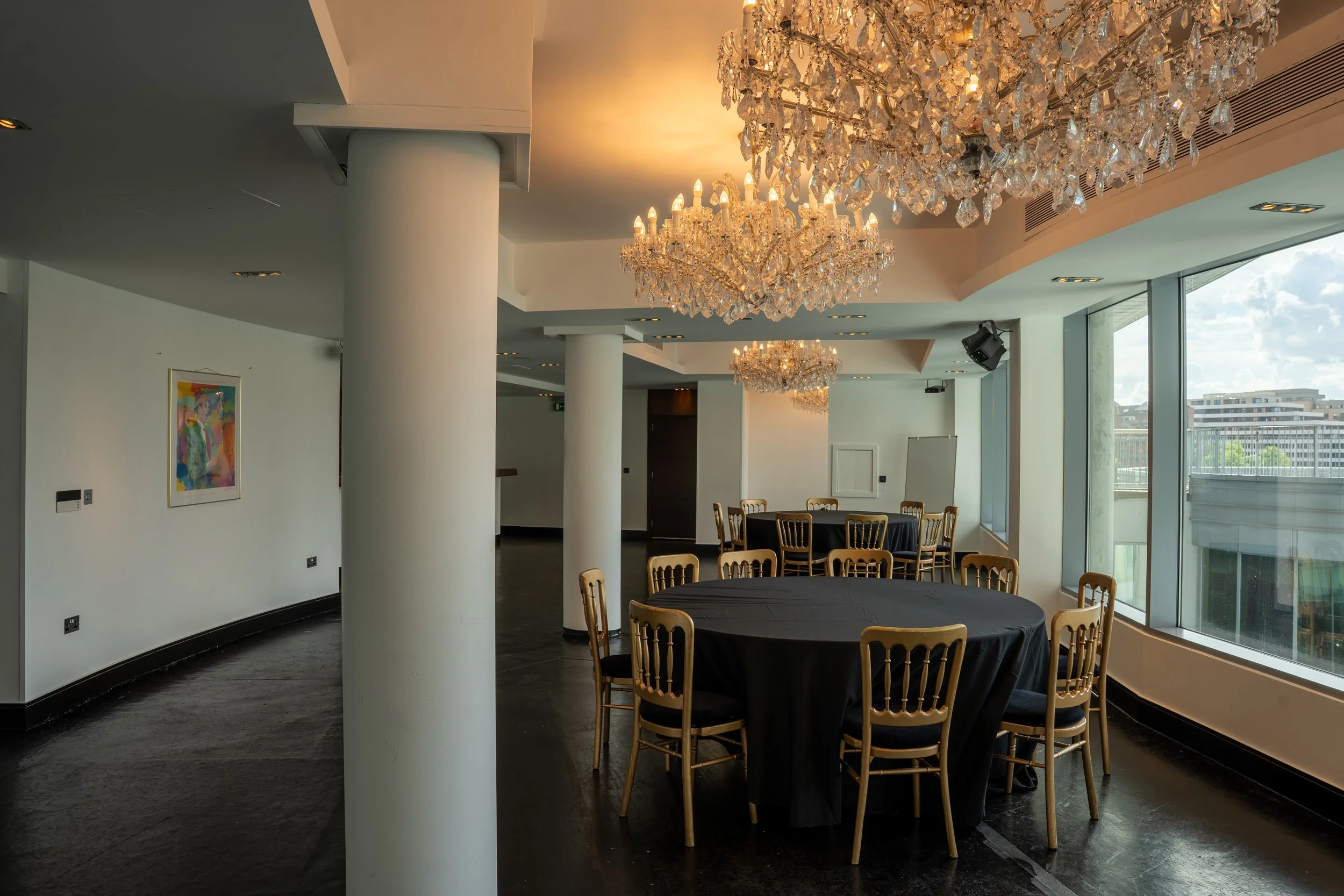 Elegant event space with multiple crystal chandeliers hanging from the ceiling, round tables covered with black tablecloths, surrounded by gold chairs, and large floor-to-ceiling windows showing city buildings and blue sky outside.