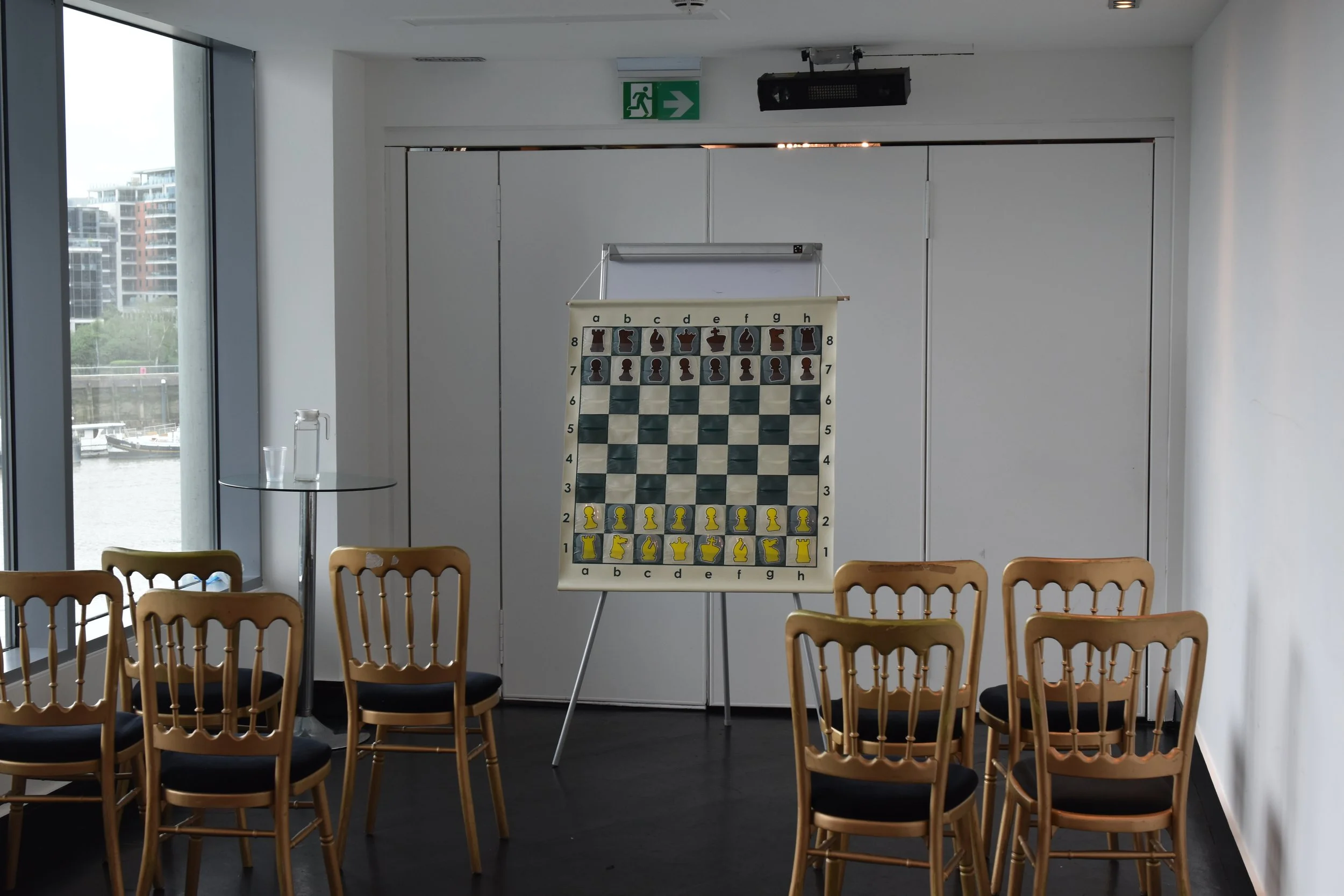 An indoor room with a large window showing a cityscape and boats on a river. There is a chessboard on a stand, with chess pieces arranged in the starting position. Several wooden chairs with black cushions are arranged in rows facing the chessboard. 