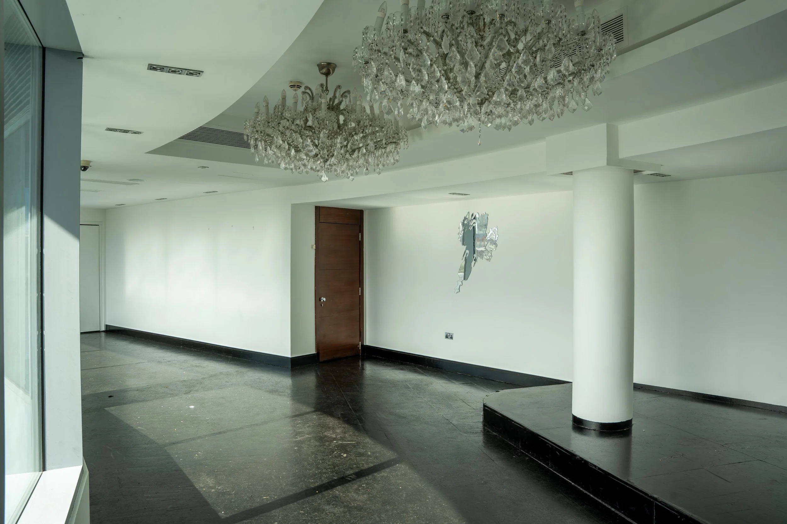 An empty modern interior space with dark tile flooring, white walls, and large crystal chandeliers hanging from the ceiling.