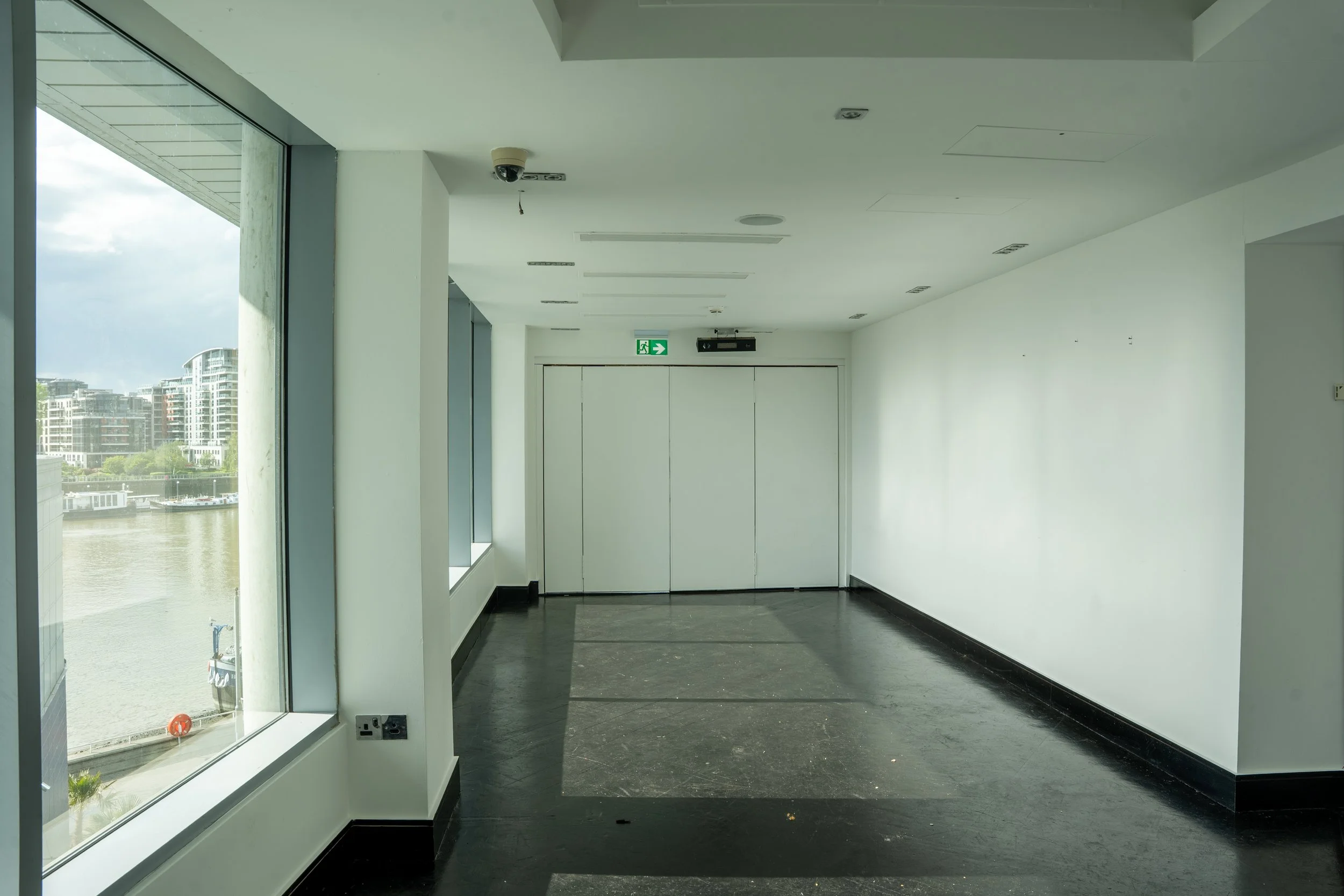 Empty indoor space with large windows revealing a cityscape and waterway outside. The room has white walls, a black floor, and an exit door with a green emergency exit sign above it.