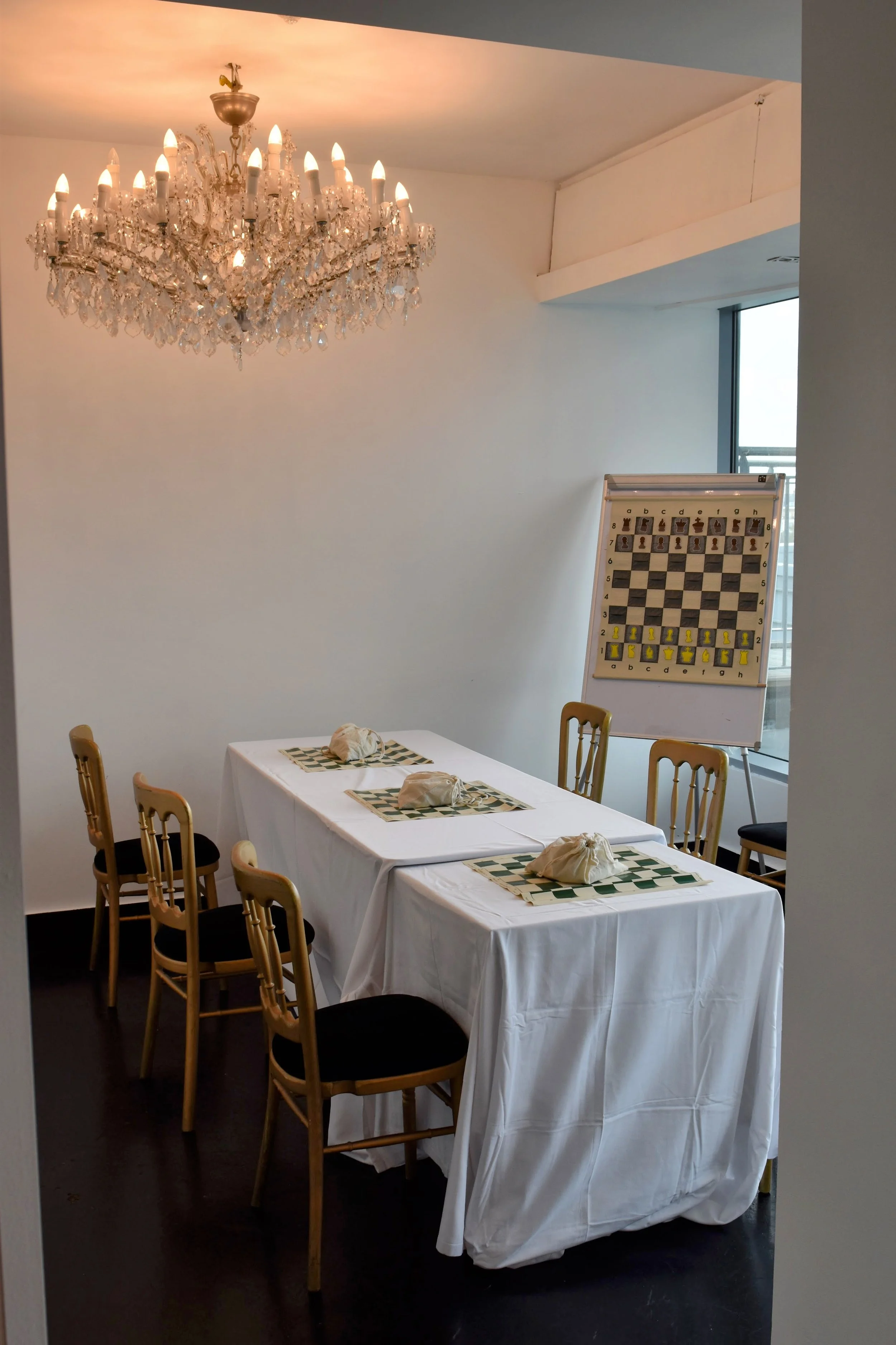 Indoor room with a large chandelier hanging from the ceiling, a table with white tablecloths and folded napkins, chessboard on an easel, and chairs around the table.