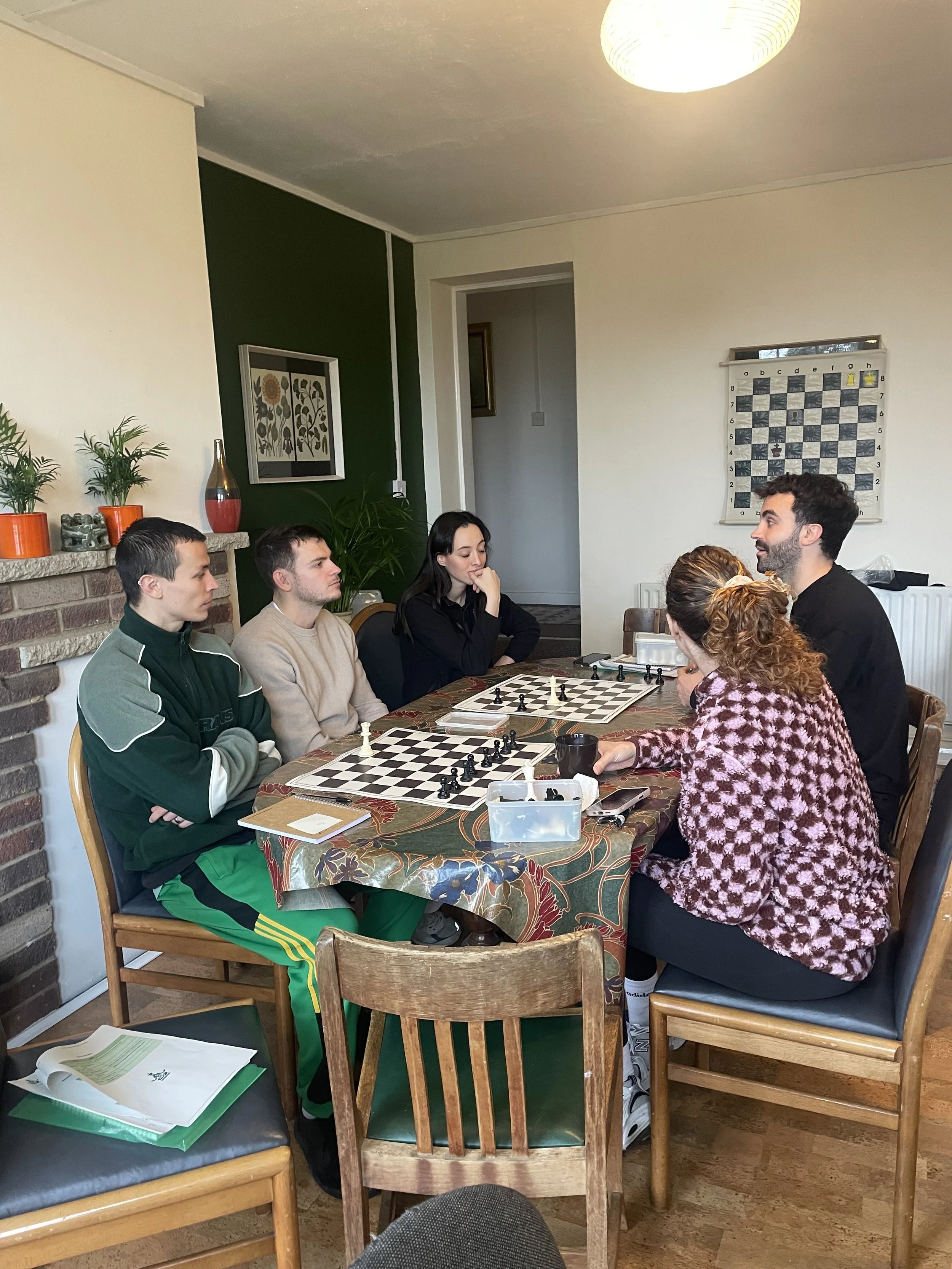 A group of six people sitting around a table playing chess in a cozy room, some with focused expressions. The room has a brick fireplace, potted plants, and a wall with artwork and a large chessboard. The table has a patterned cloth, and there are ch