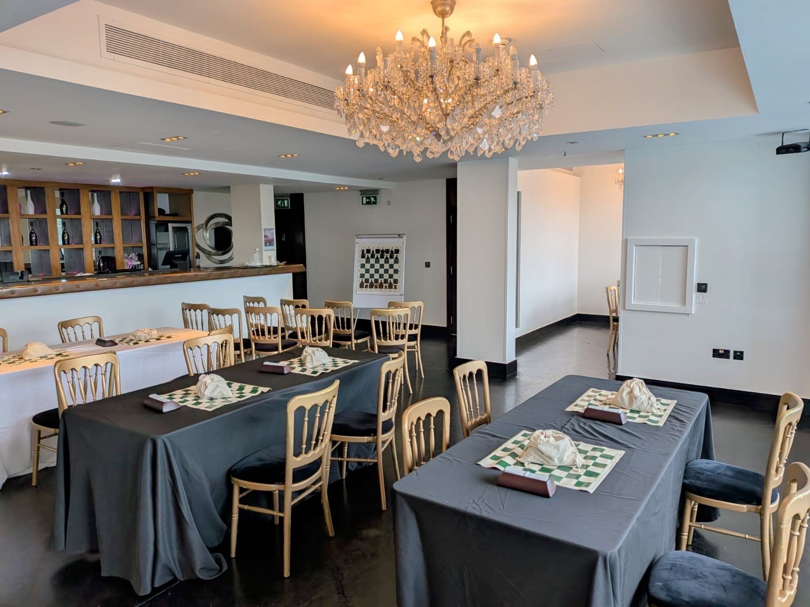 Interior of a restaurant dining room with tables covered in gray tablecloths, set with napkins and silverware, wooden chairs, a chandelier hanging from the ceiling, a bar area with shelves and bottles, and a wall with a decorative mirror.
