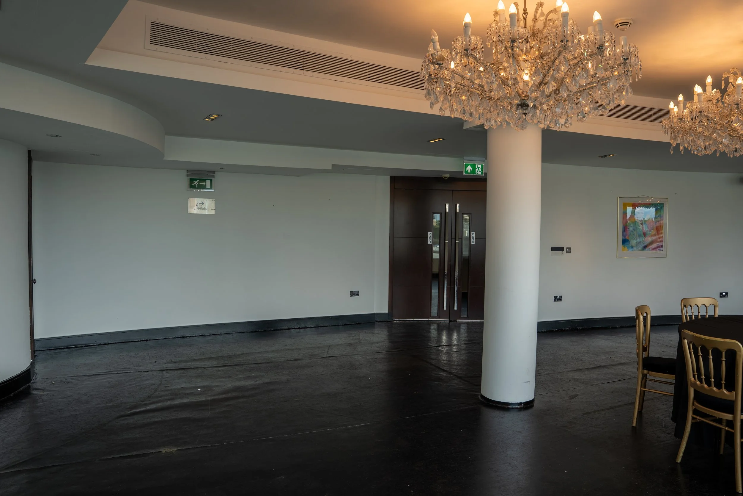 Interior of a room with dark hardwood floors, white walls, and a white pillar. Two large crystal chandeliers hanging from the ceiling. A framed colorful artwork on the wall, exit signs above doors, and wooden chairs around a table.